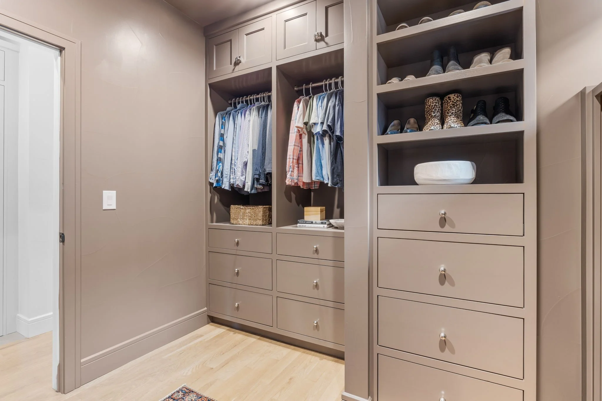 Walk-in closet with organized clothing and shoes storage, featuring hanging shirts, woven basket, books, and neatly arranged shoes on shelves.