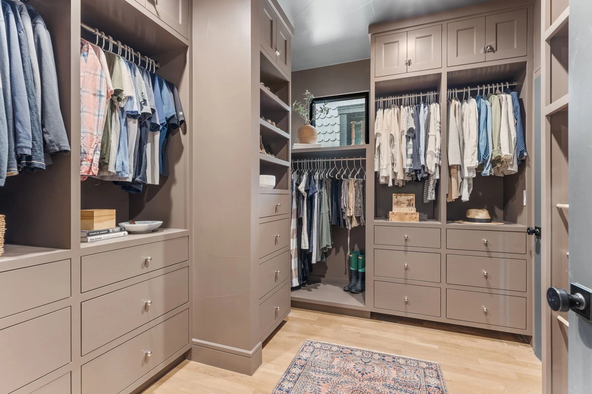 A walk-in closet with beige cabinetry and clothes hanging on rods, a small window, a plant, shelves, and drawers, with a wooden floor and a small rug.
