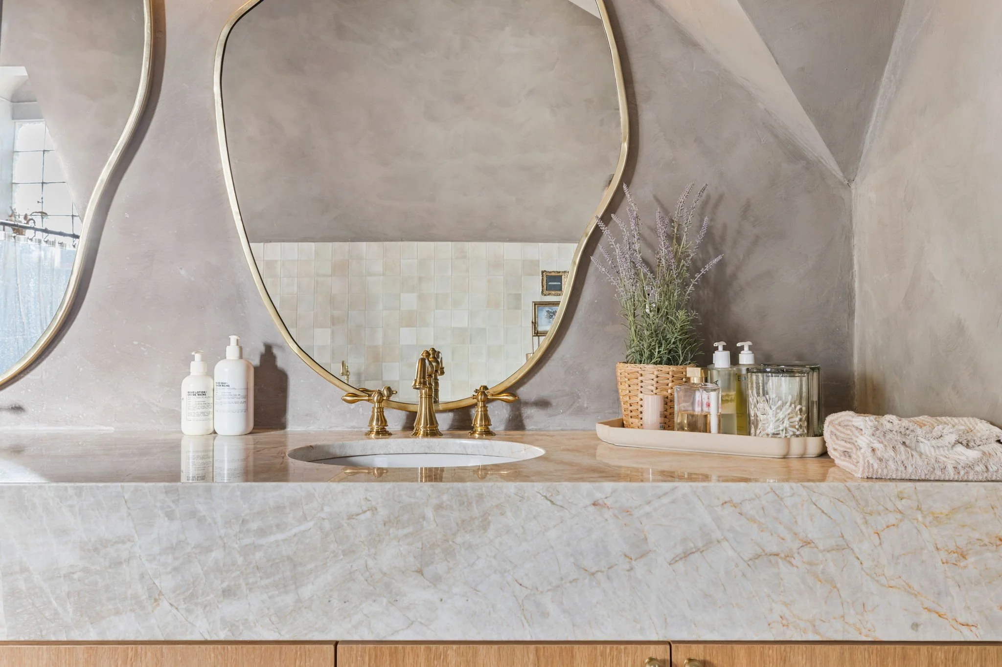 Bathroom vanity with marble top, large mirror with gold frame, and various bottles of skincare or soap, a pot of lavender, a bamboo basket, and folded cloth on the right side.
