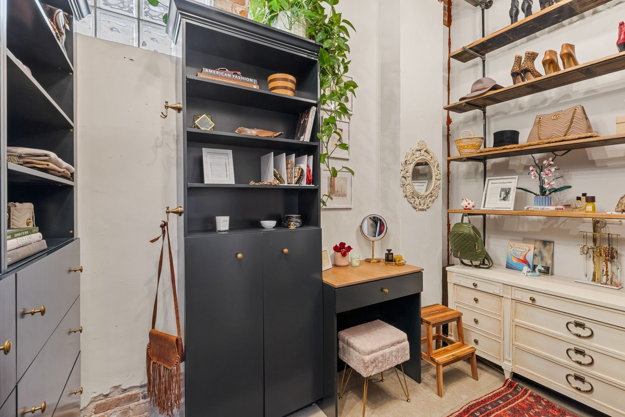 Interior of a boutique or home decor store with shelves displaying handbags, shoes, and accessories, a black cabinet, a white dresser, and decorative items like a mirror, flowers, and books.