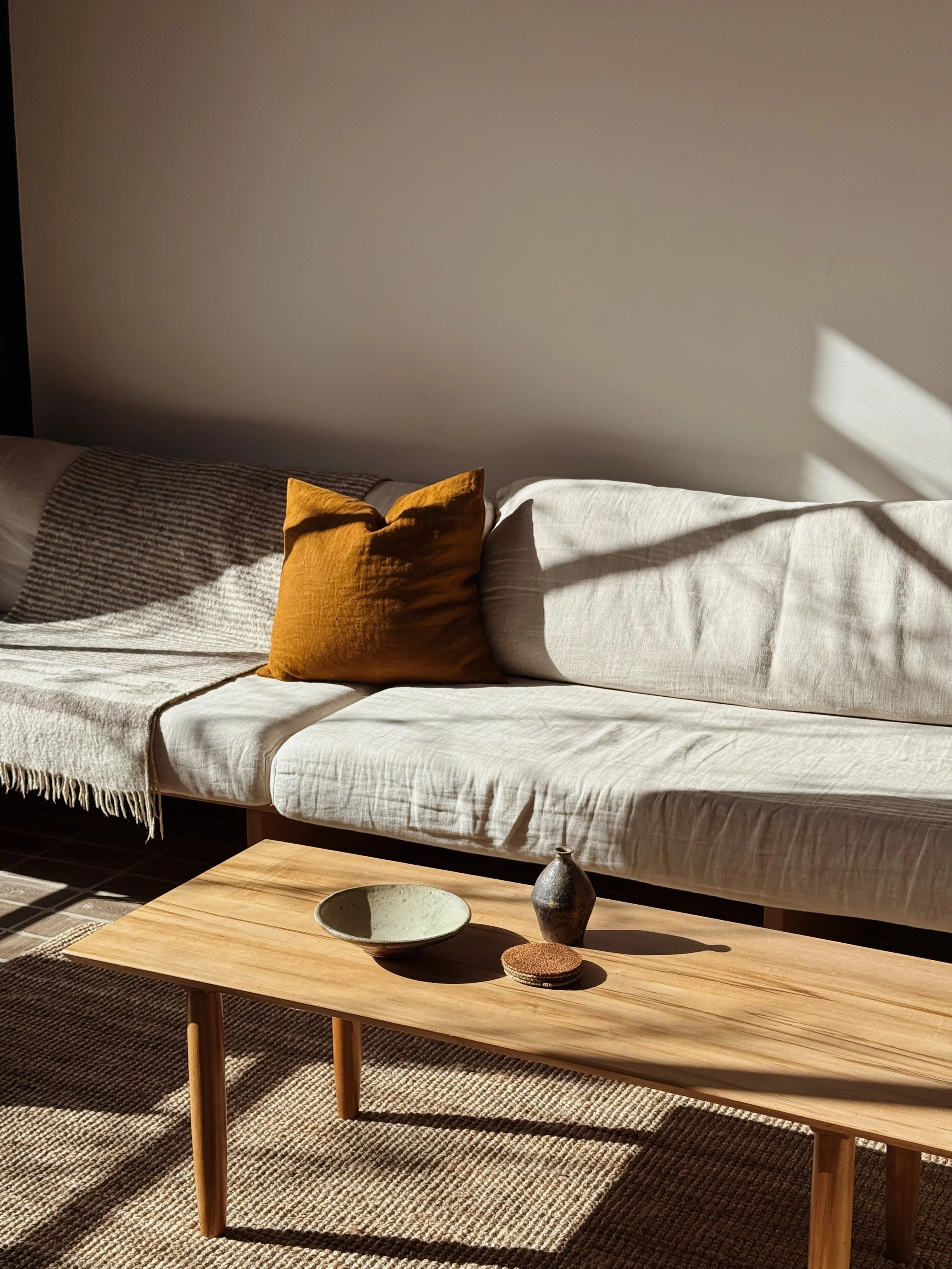 Living room with a white sofa, a mustard-yellow pillow, a beige throw blanket, a wooden coffee table with ceramic bowl, small black vase, and coaster, beige rug, and sunlight casting shadows on the sofa and wall.