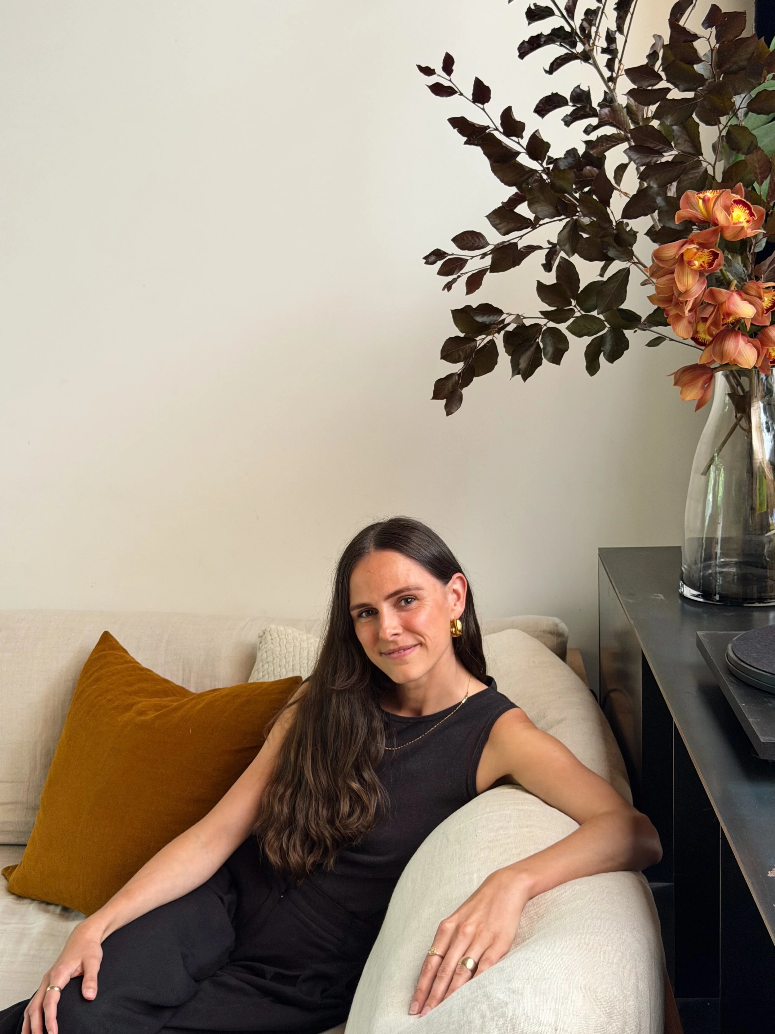 A woman with long brown hair, wearing a black sleeveless top, gold jewelry, and earrings, sitting on a cream-colored couch with a brown cushion. She is smiling slightly while resting her left arm on the armrest and her right arm on her lap. Behind her, there is a black console table with a large glass vase holding dark purple leaves and pinkish-orange flowers. The background is a plain off-white wall.