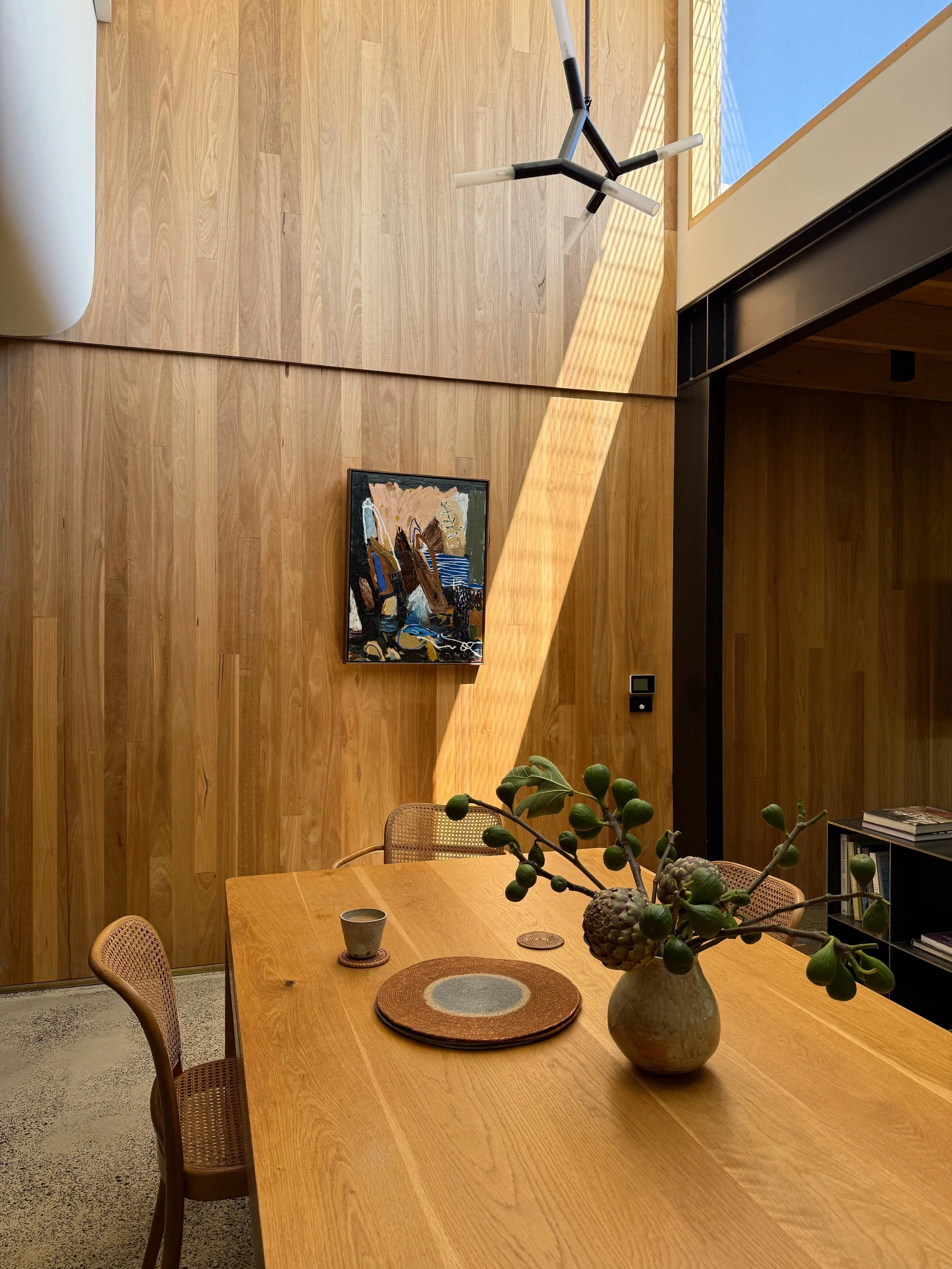 A dining room with a wooden table, rattan chairs, and a vase with branches and large seed pods. There is a framed abstract painting on a wooden wall, and a modern black and white geometric light fixture hanging from the ceiling.