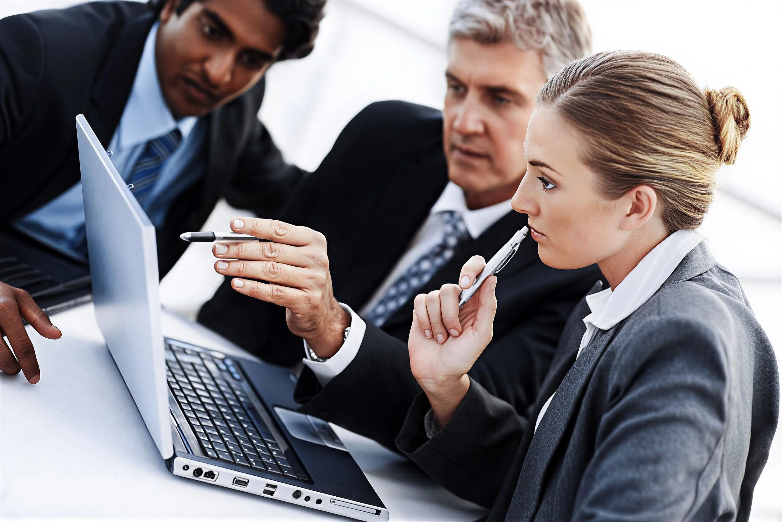 Three business professionals in suits discussing around a laptop, with one woman holding a pen near her face.