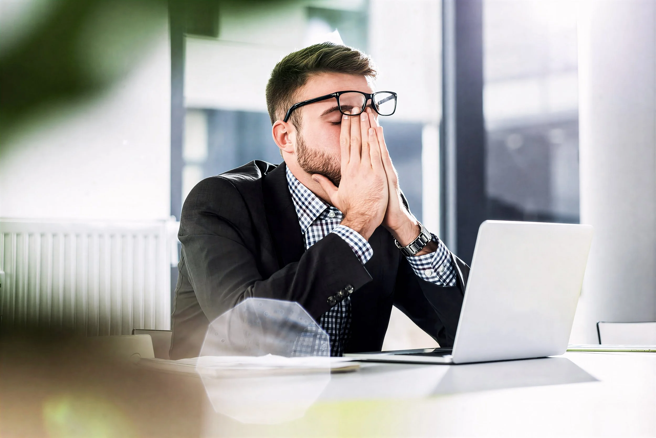 Frustrated man in business attire sitting at a desk with a laptop, holding his face and eyes closed in disappointment