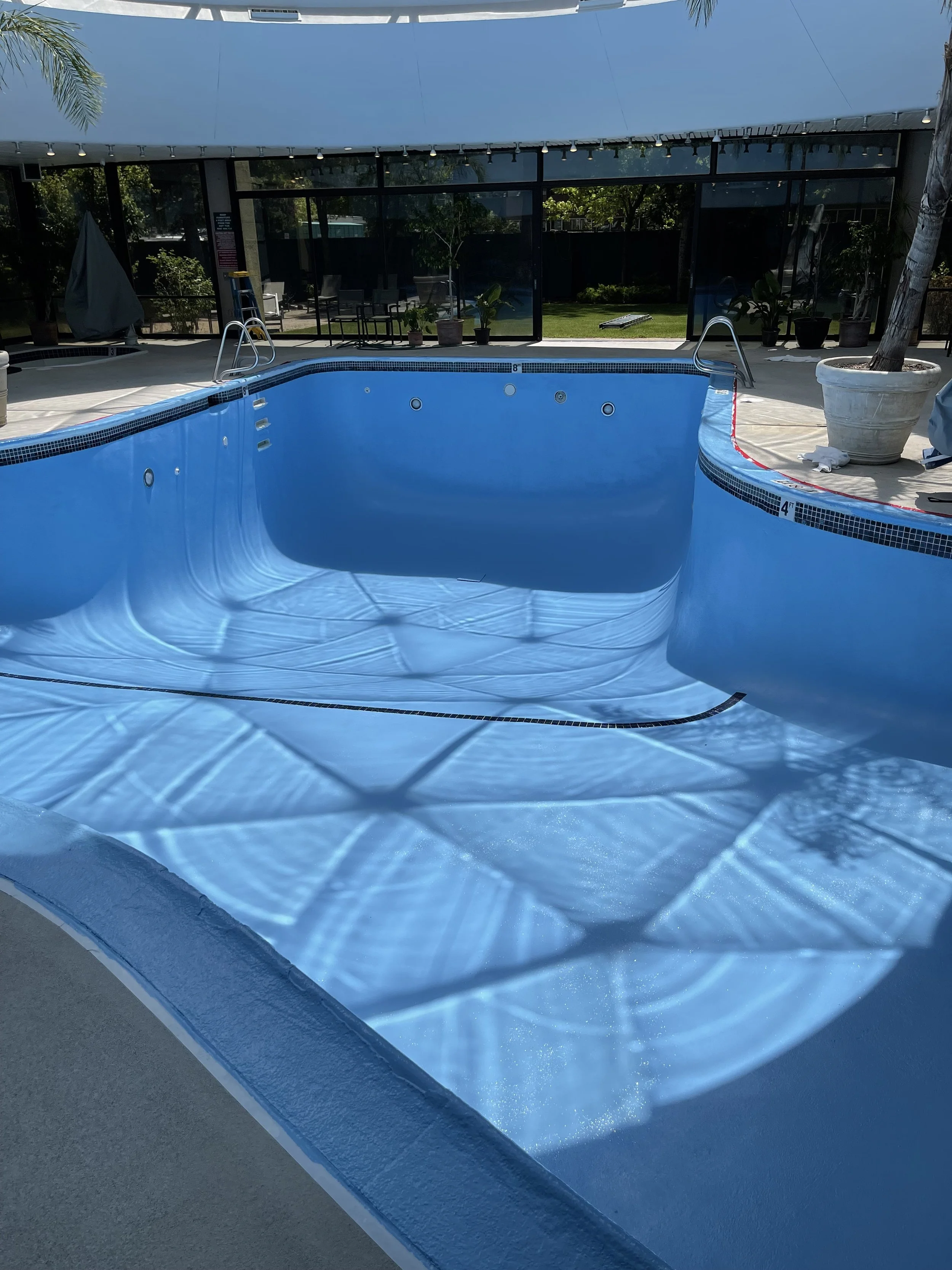 Empty swimming pool with shadows of palm trees on the water surface, surrounded by patio area with potted plants and outdoor furniture.