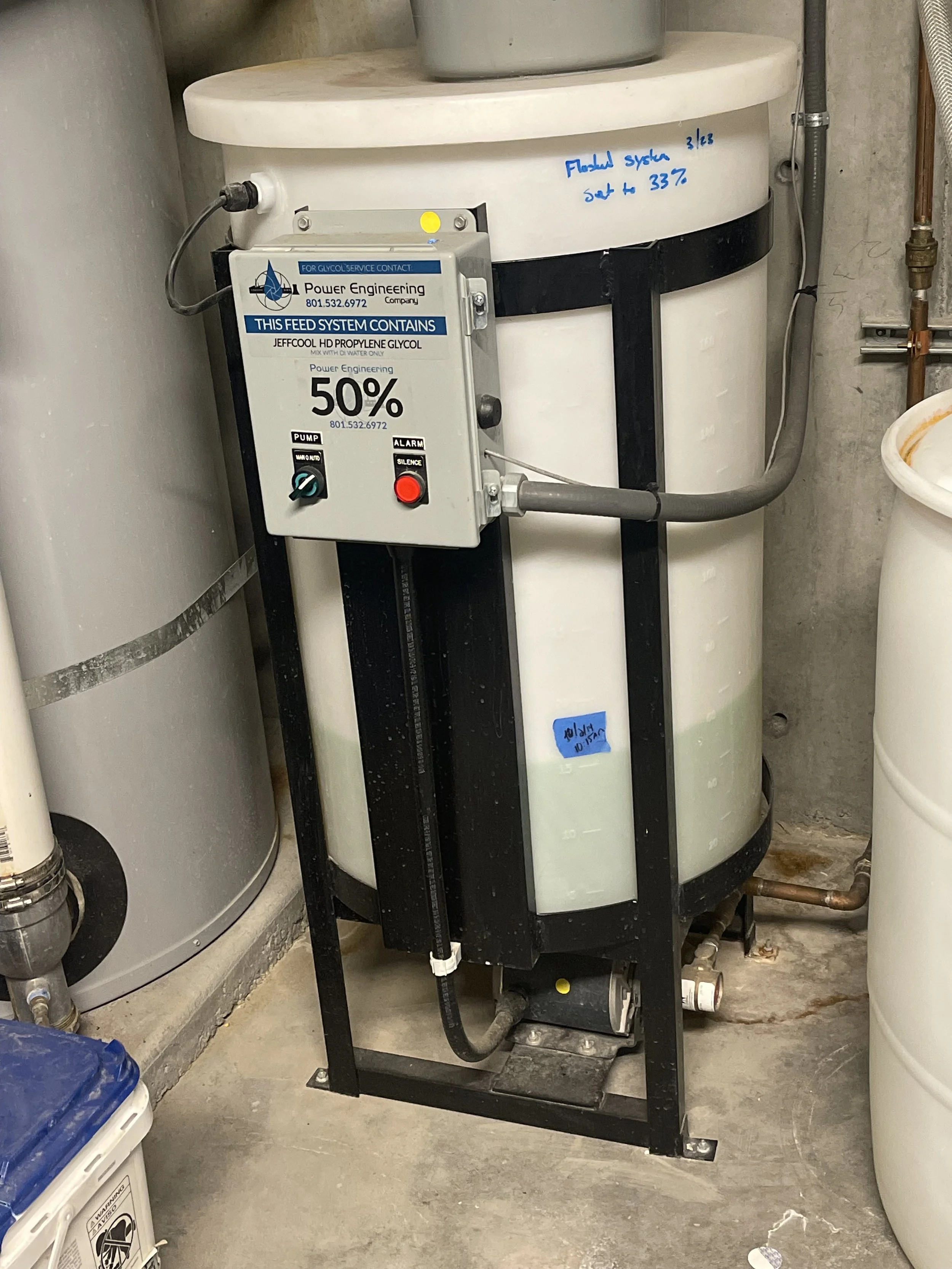 A glycell feed system containing 50% glycol solution, mounted on a black metal stand in a utility room with concrete floor and surrounding equipment.