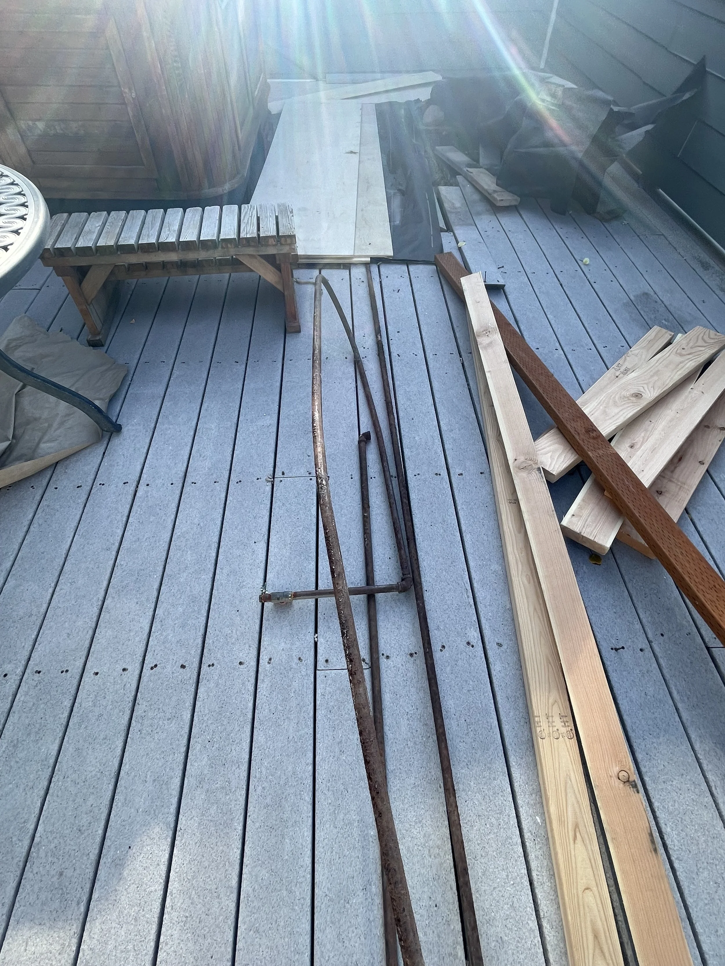 Construction tools and wood planks on a wooden deck, with a bench and a trash bag nearby.