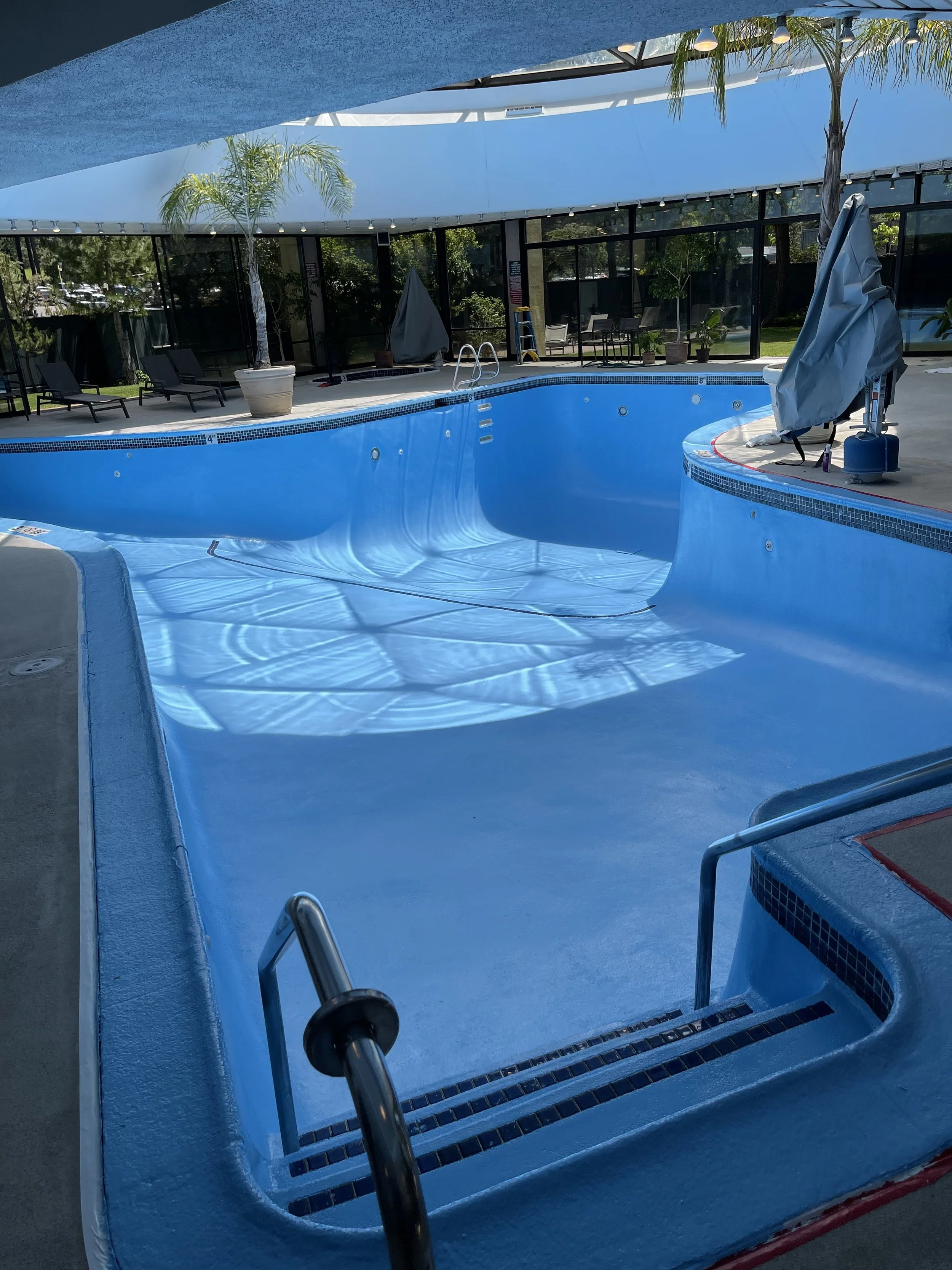 Empty indoor swimming pool with blue lining, surrounded by lounge chairs, potted plants, and large windows with natural light.