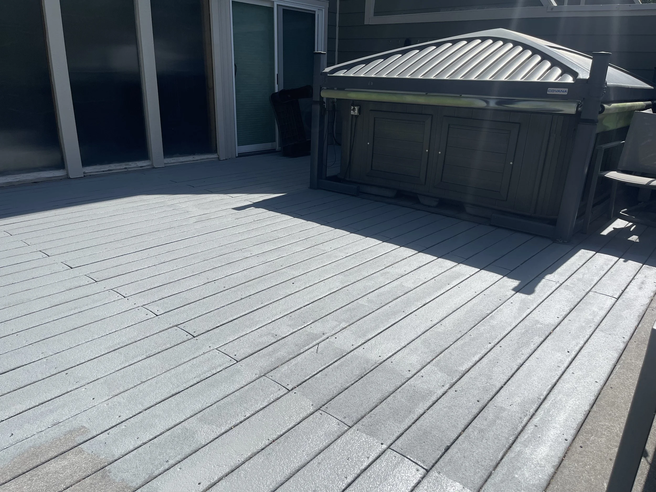 Empty outdoor patio with light gray wooden floorboards, sliding glass door, black hot tub, and black outdoor chair.