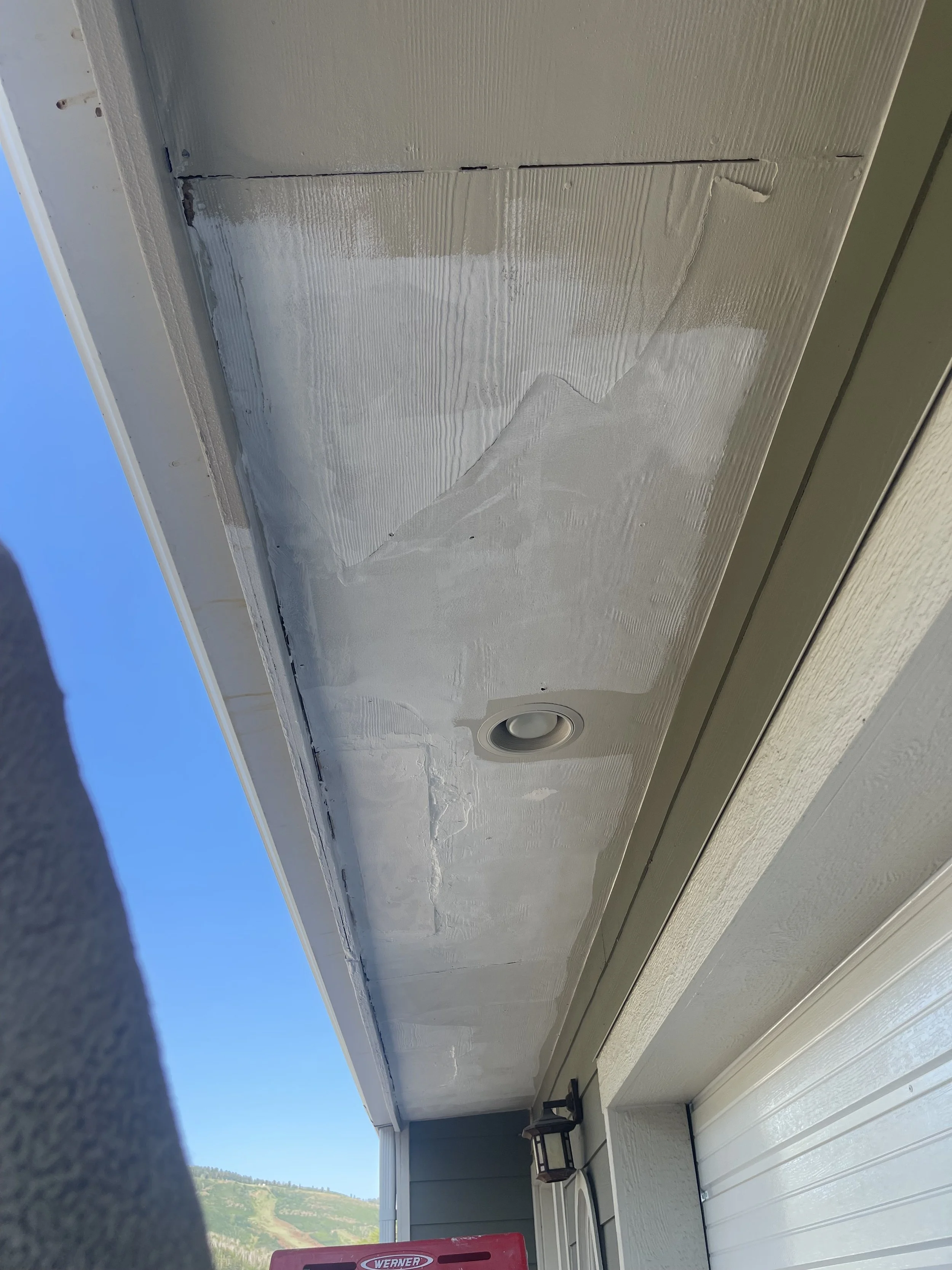 Ceiling of a house with ongoing painting work, a recessed light fixture, and mountain scenery visible outside through part of the house structure.