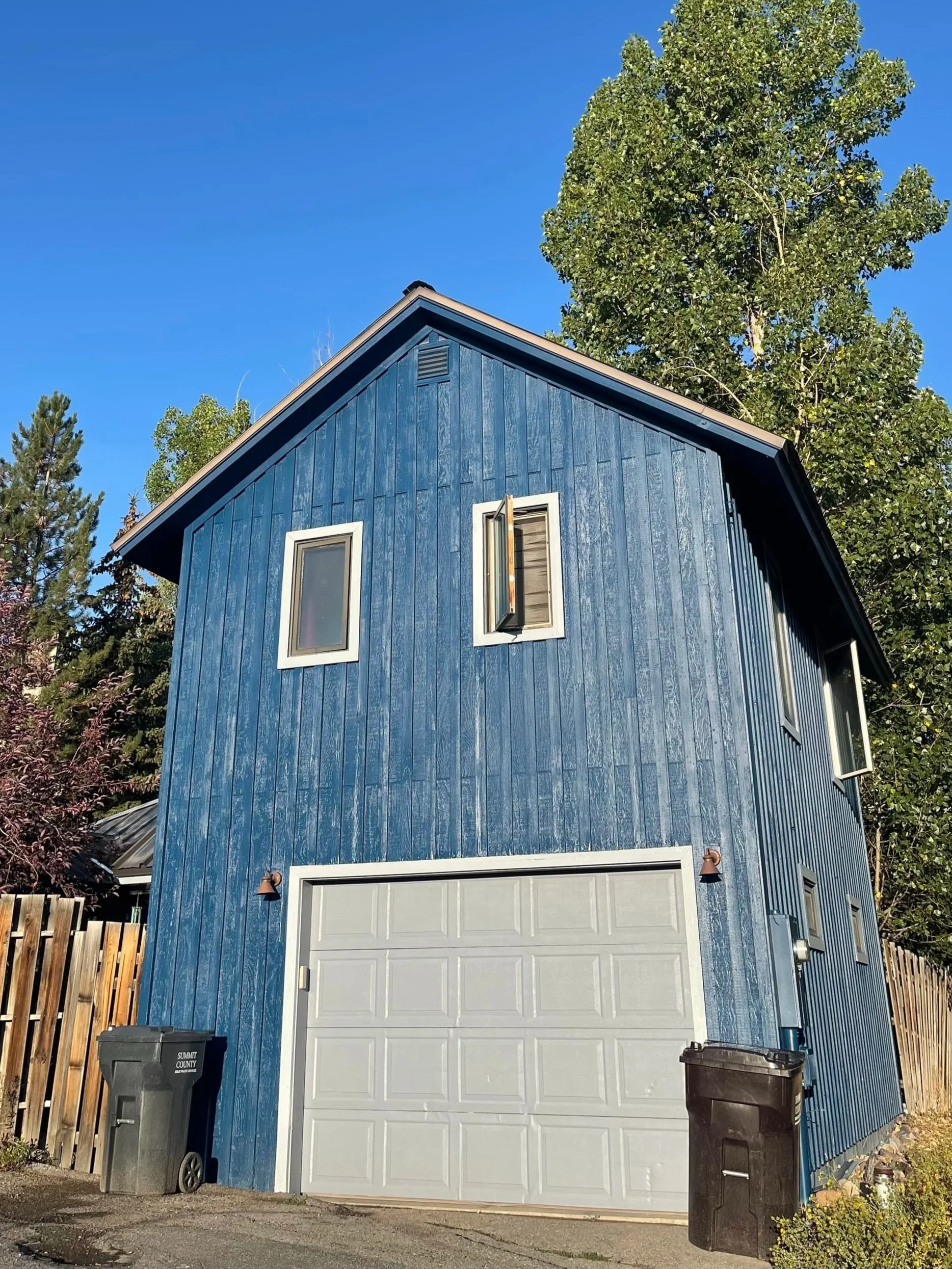 A blue two-story house with a garage door on the front, surrounded by tall trees and a wooden fence, set against a clear blue sky.