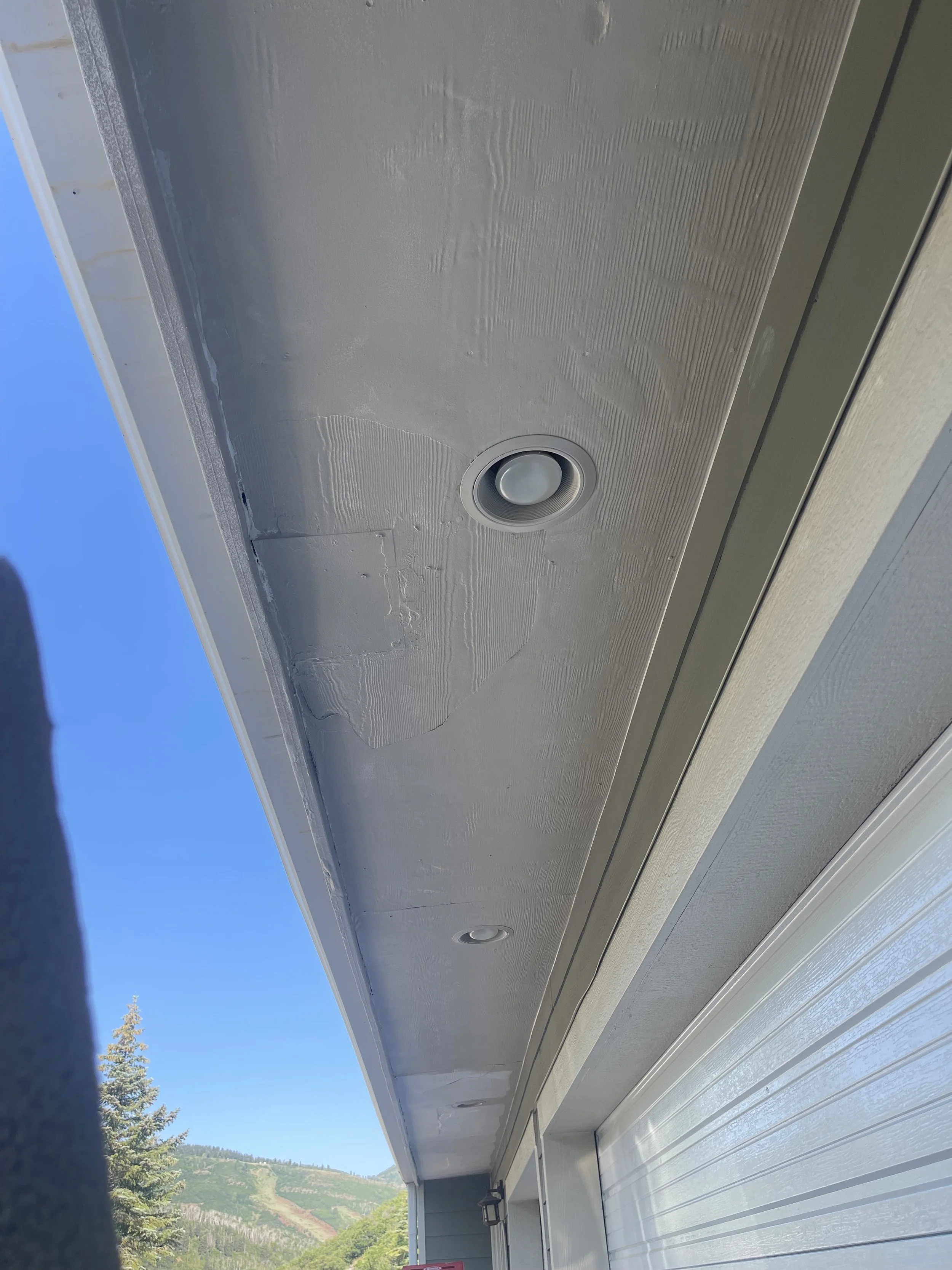 Close-up of a house's exterior ceiling with an installed recessed light fixture, showing textured white wall surface and part of a white garage door, with mountains and trees in the background.
