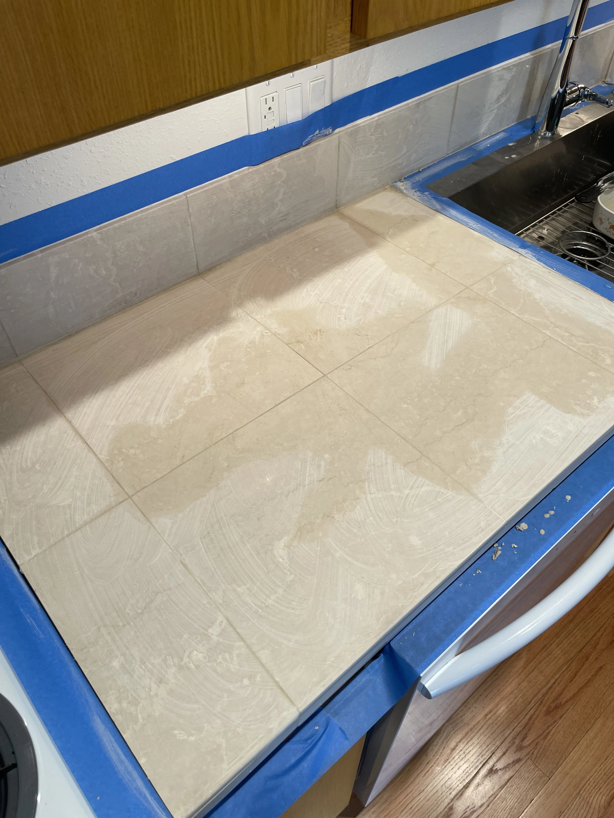 Kitchen countertop with tiles being installed, blue painter's tape along the edges, a sink on the right, and a wall outlet in the background.
