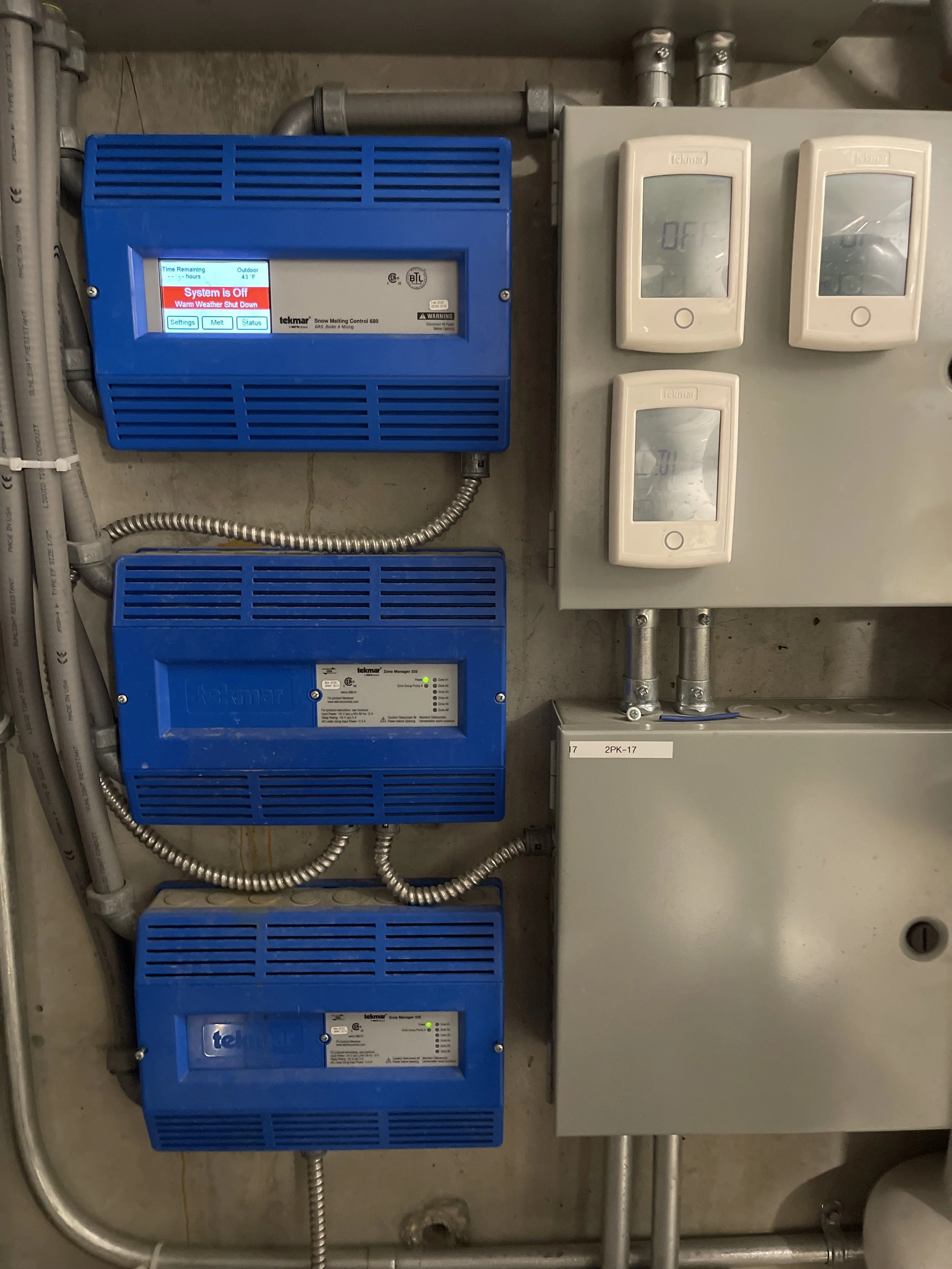 Electrical control panel with three blue Tekmar snow melting system controllers and three white thermostats on a gray wall.
