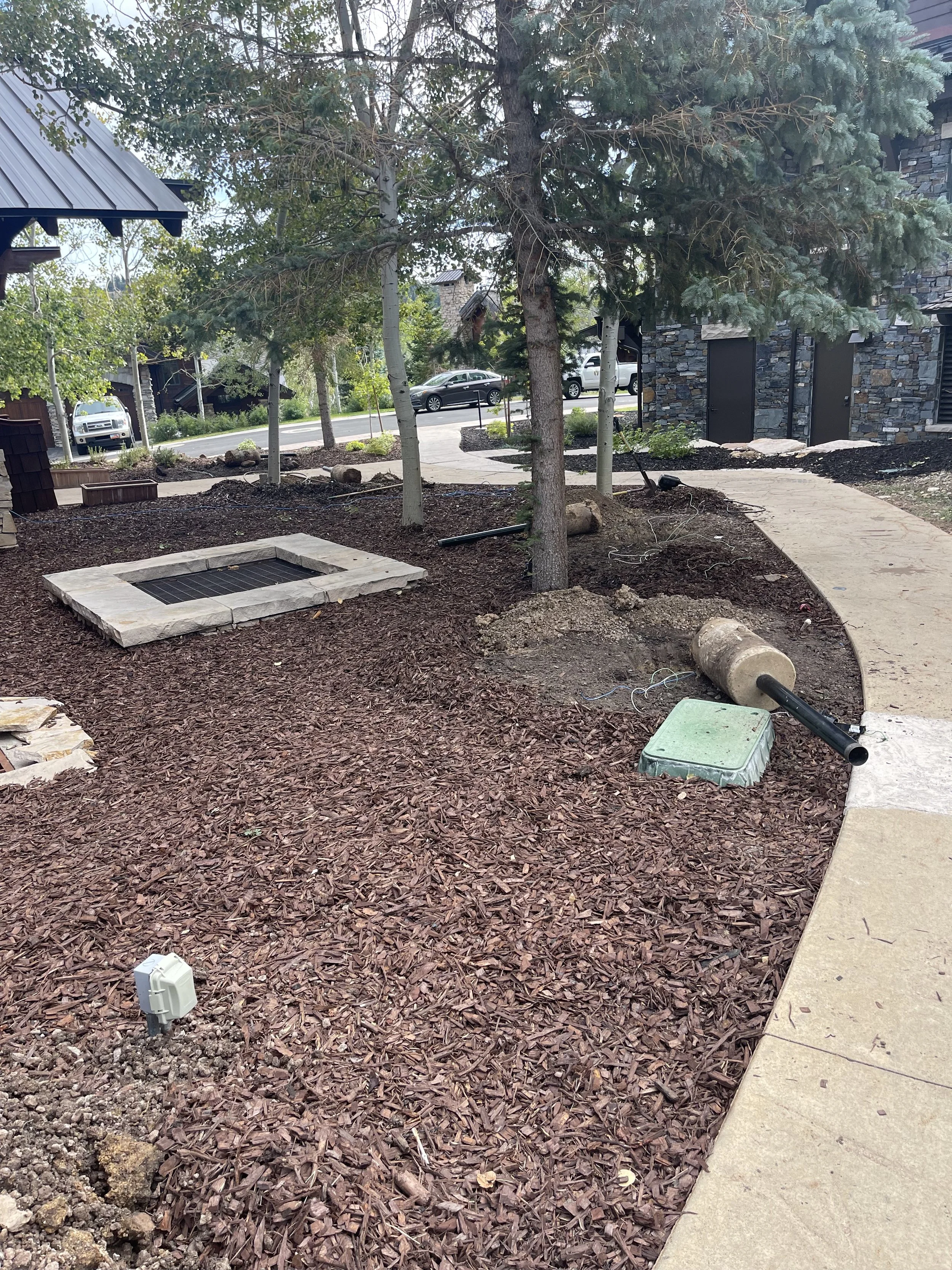 Landscape scene of a landscaping project with mulch, trees, a stone-lined square feature, a green utility box, and pipes on a pathway, with parked cars and a building in the background.