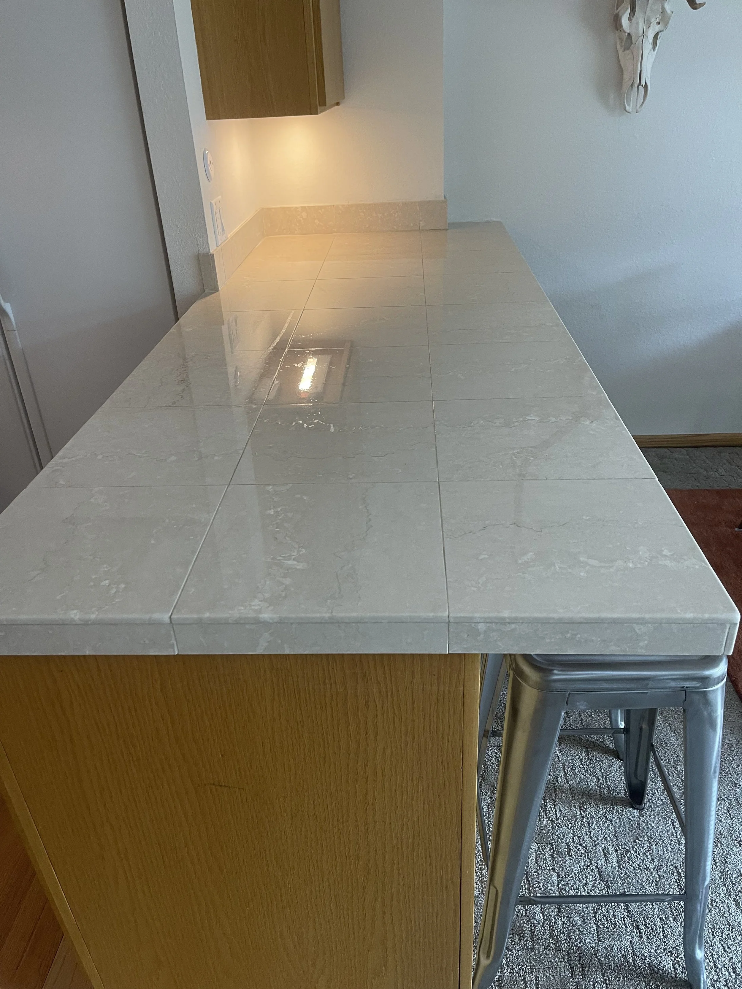 A kitchen counter with a light-colored tiled surface, a metal stool tucked underneath, and a white skull décor on the wall to the right.