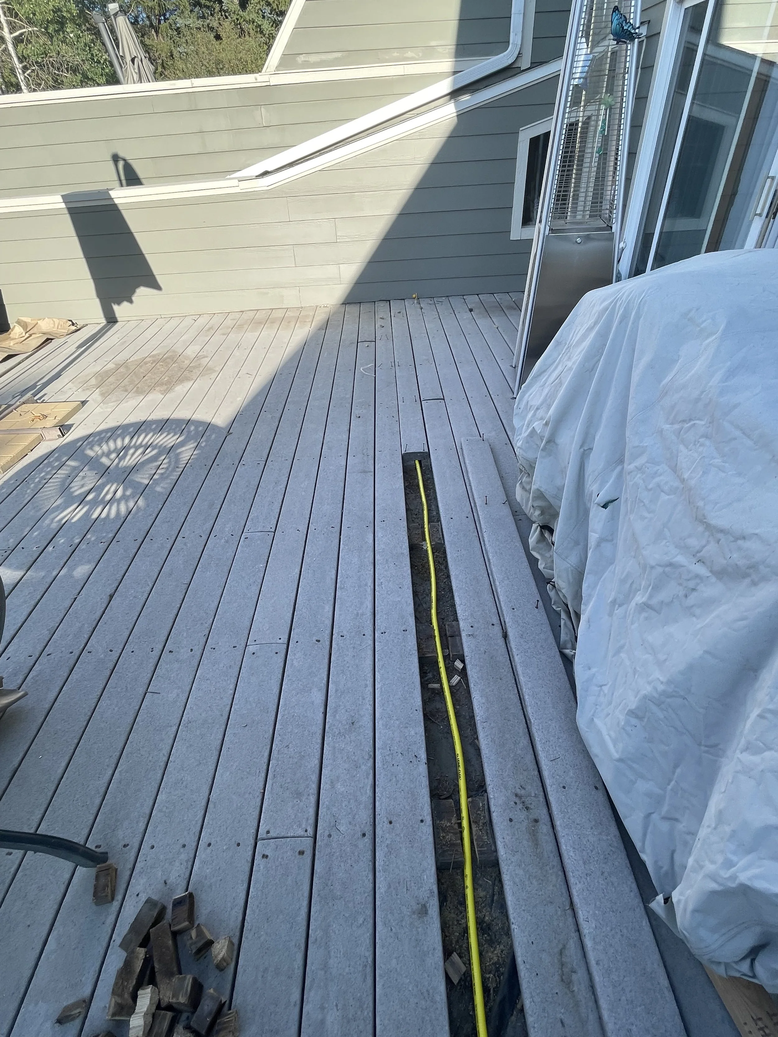 Construction work on a wooden deck showing a long rectangular cutout with yellow electrical wire running through it. Some tools and wood pieces are scattered on the deck, and a covered object is on the right side.