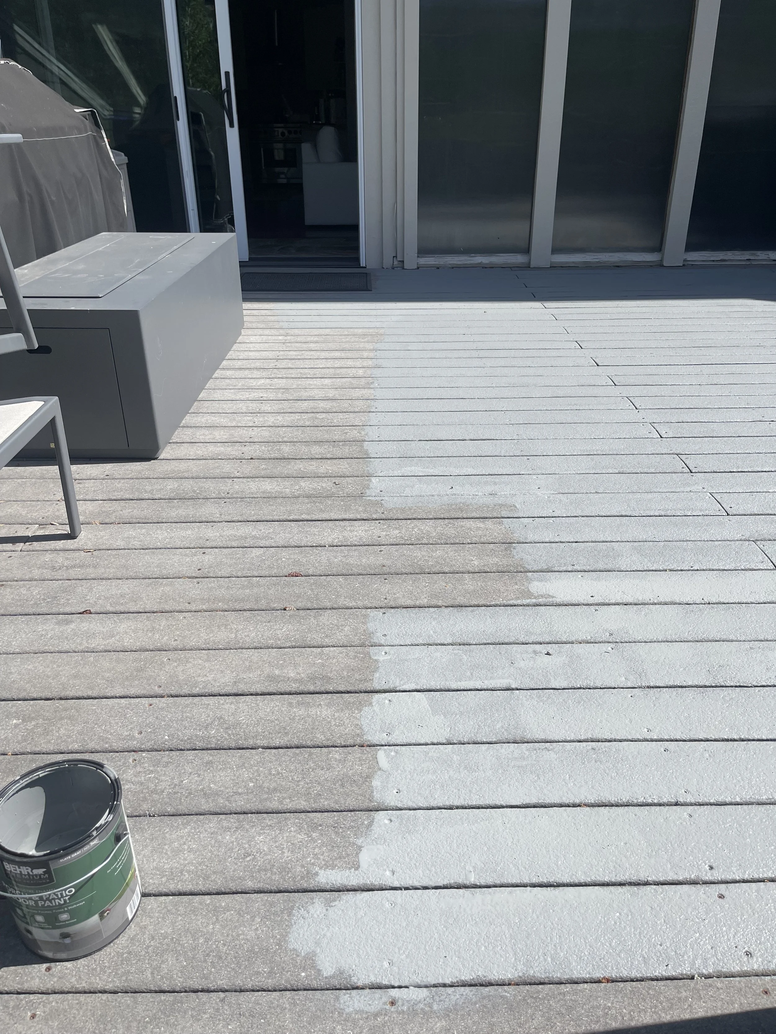 A wooden deck partially painted grey, with paint and paint can visible, outside a house with sliding glass door.