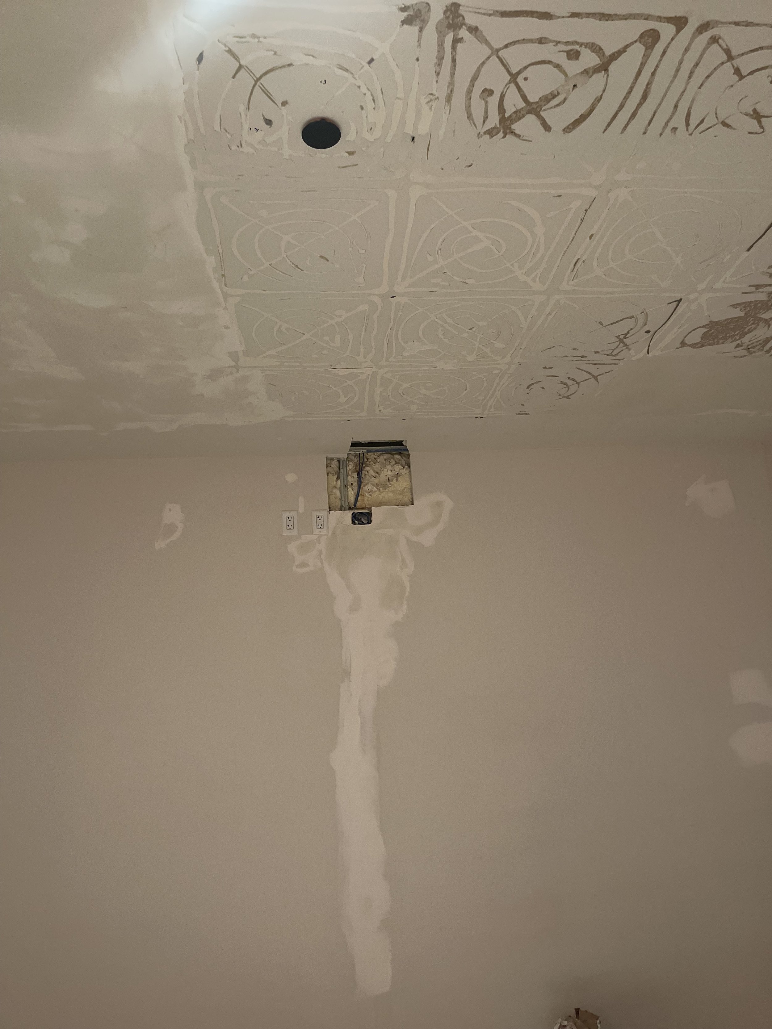 A room under construction with a partially finished ceiling showing tape and drywall mud, a hole in the ceiling for a light fixture, and an open wall cavity with wires and plumbing visible.
