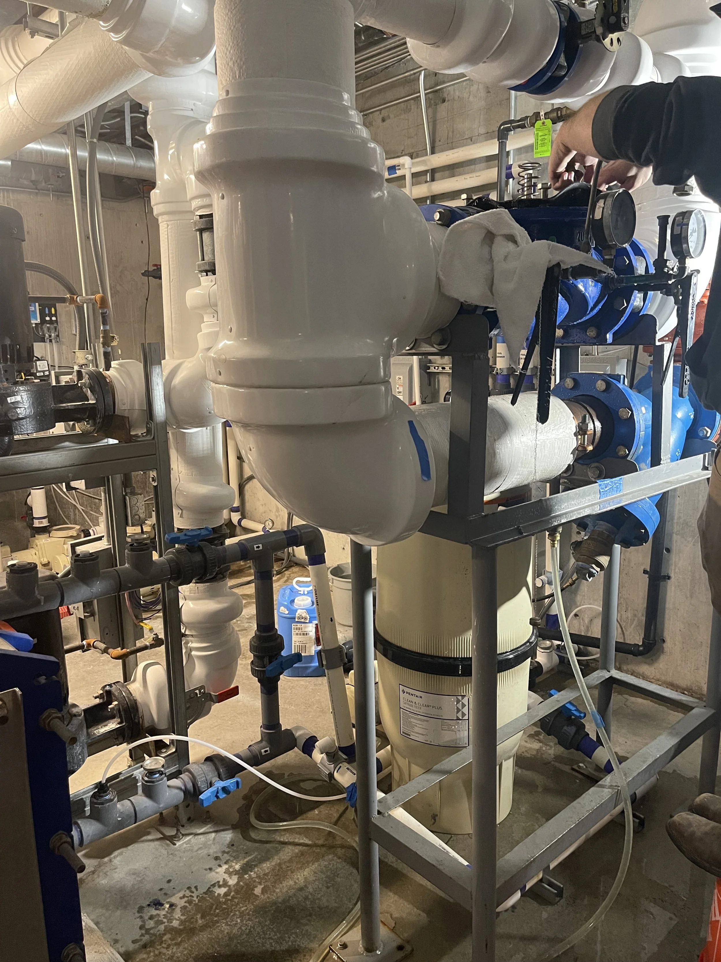 A person working on plumbing equipment in a mechanical room, installing or repairing a large filter system with various pipes and valves.