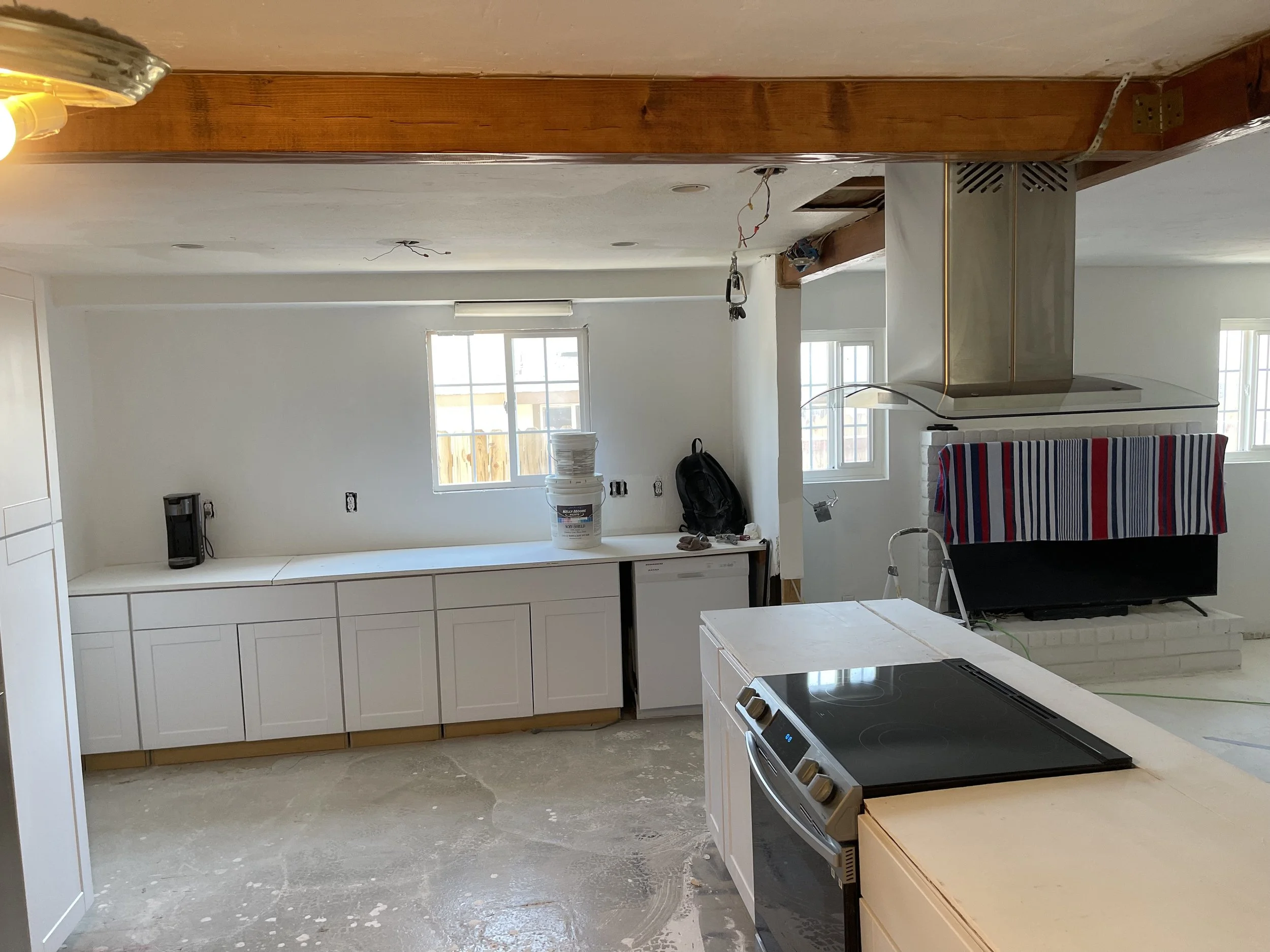 Kitchen under renovation with white cabinets, a stove on the island, and a brick fireplace above which a strip of striped fabric is draped. Construction materials and tools are visible.