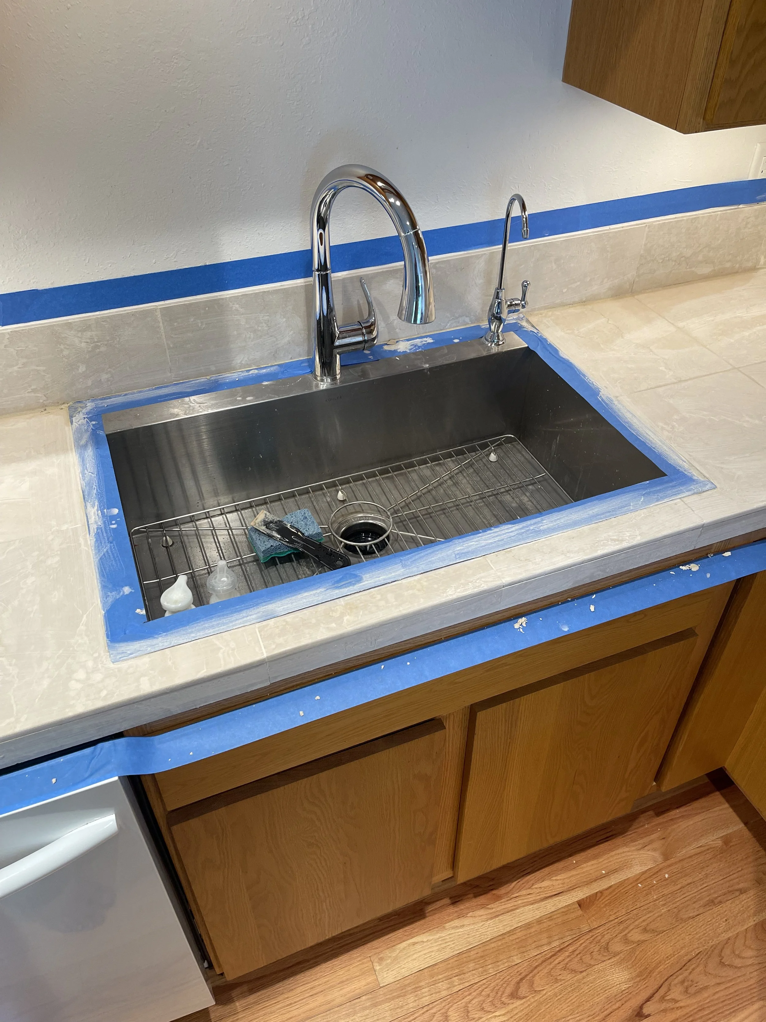 Kitchen sink with blue painter's tape around the edges of the countertop, a sponge, and other cleaning tools inside the sink. The counter and backsplash are partially taped for installation or painting.