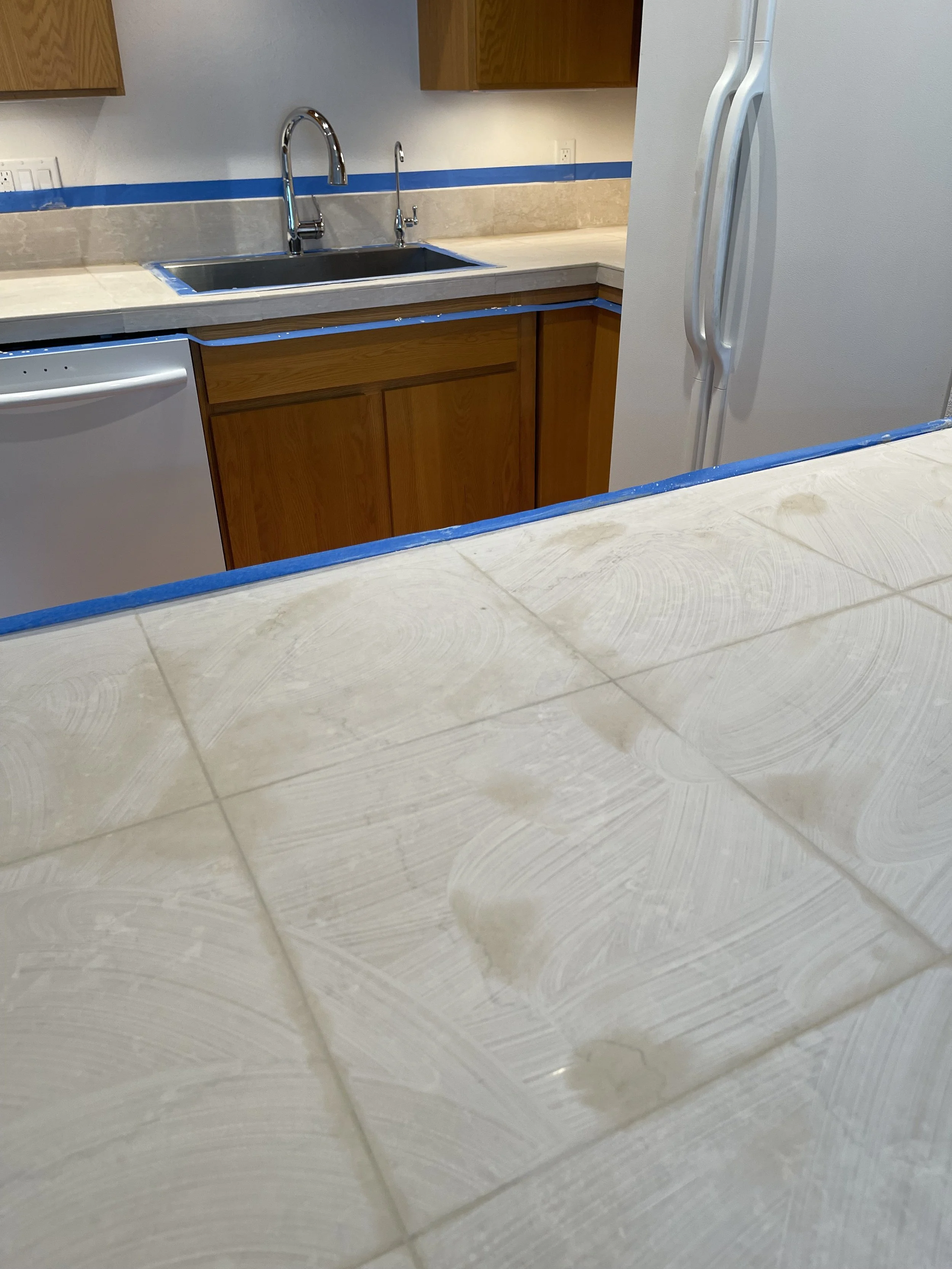 Kitchen with wooden cabinets, a double sink, a white refrigerator, and a tiled countertop with blue painter's tape.