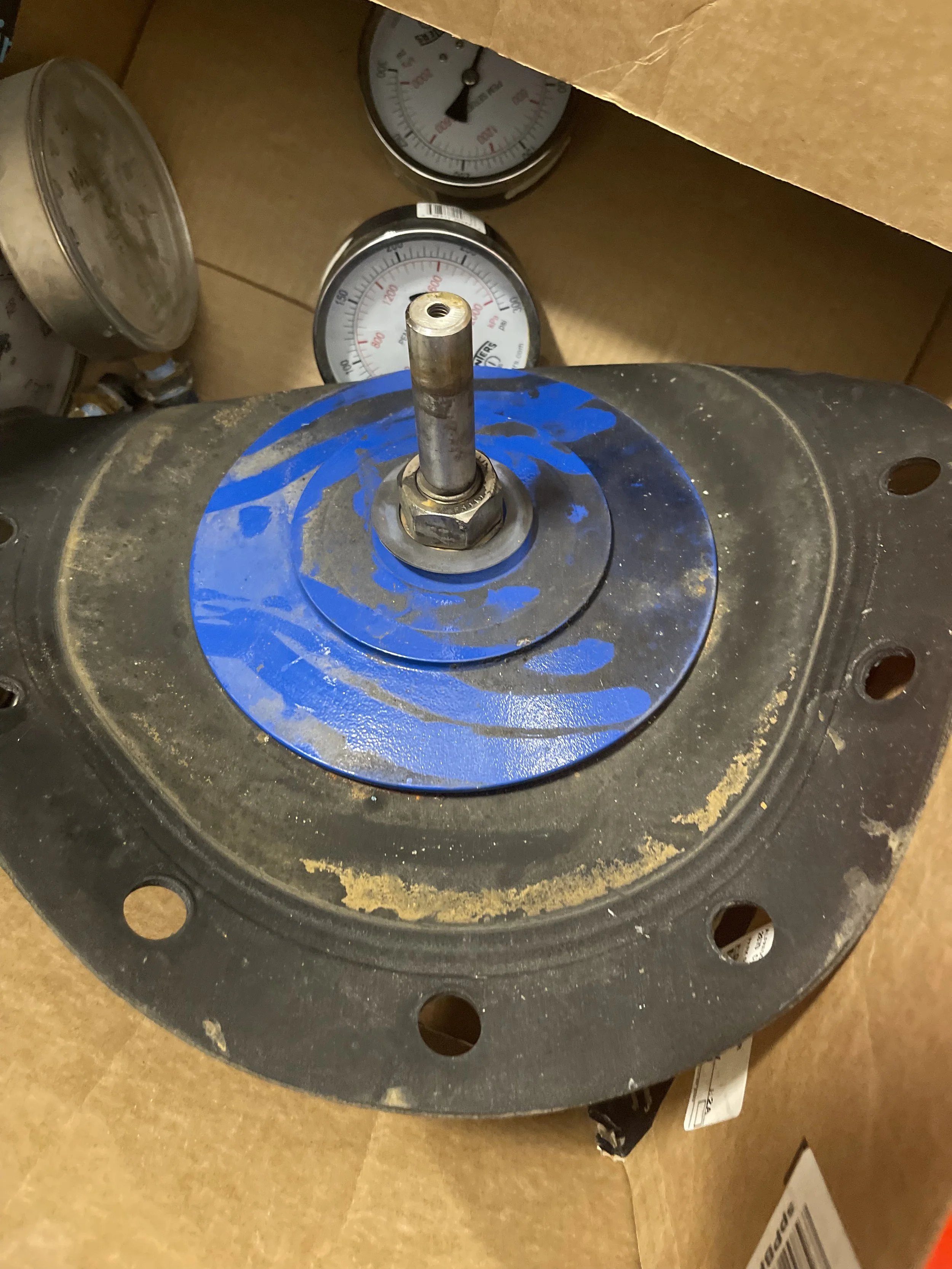 Mechanical part with a central bolt mounted on a black circular base with blue paint and rust, inside a cardboard box with pressure gauges in the background.
