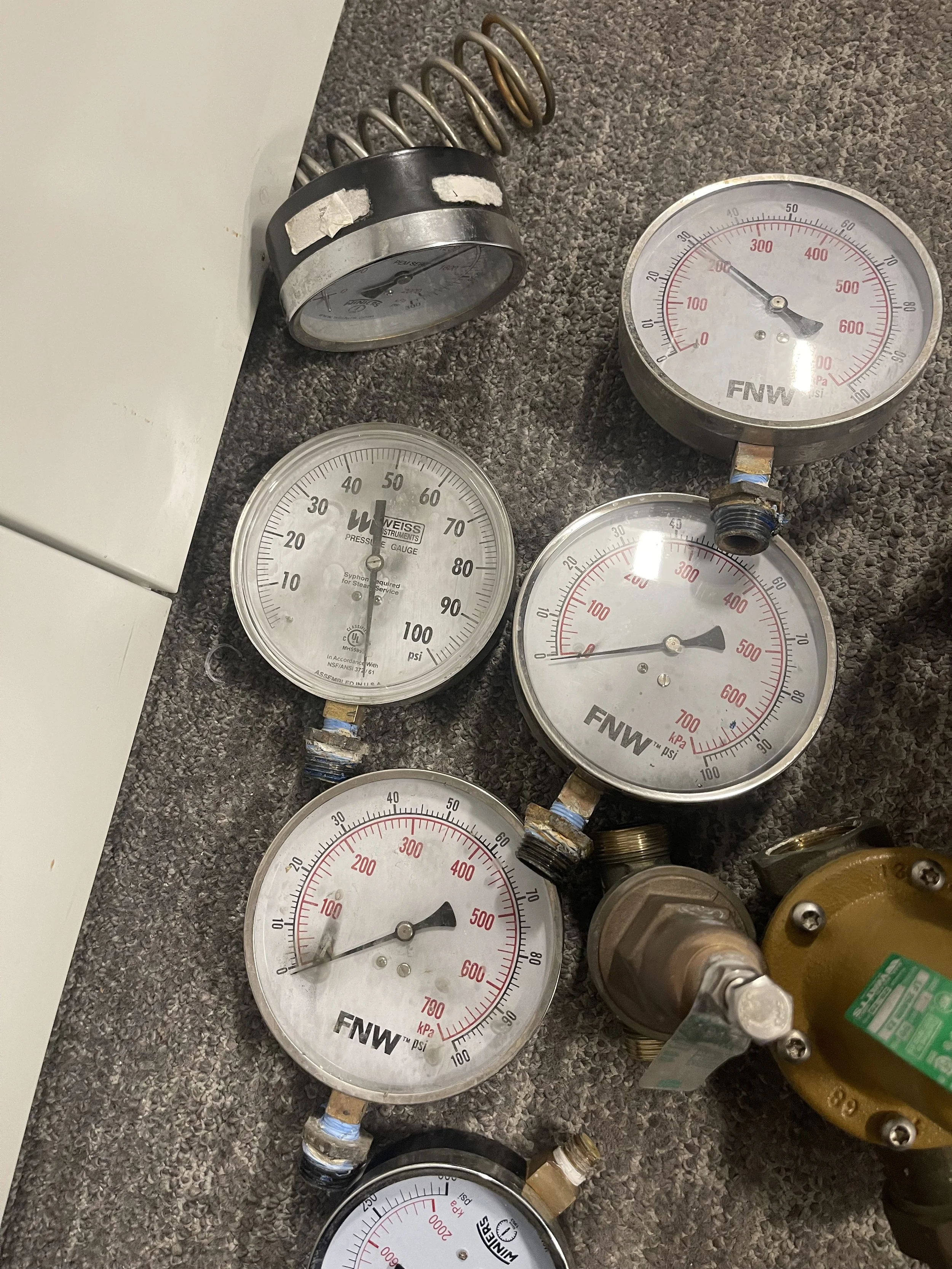 Various pressure gauges and a spring, some attached to pipes, with other gauges lying on a carpeted floor.