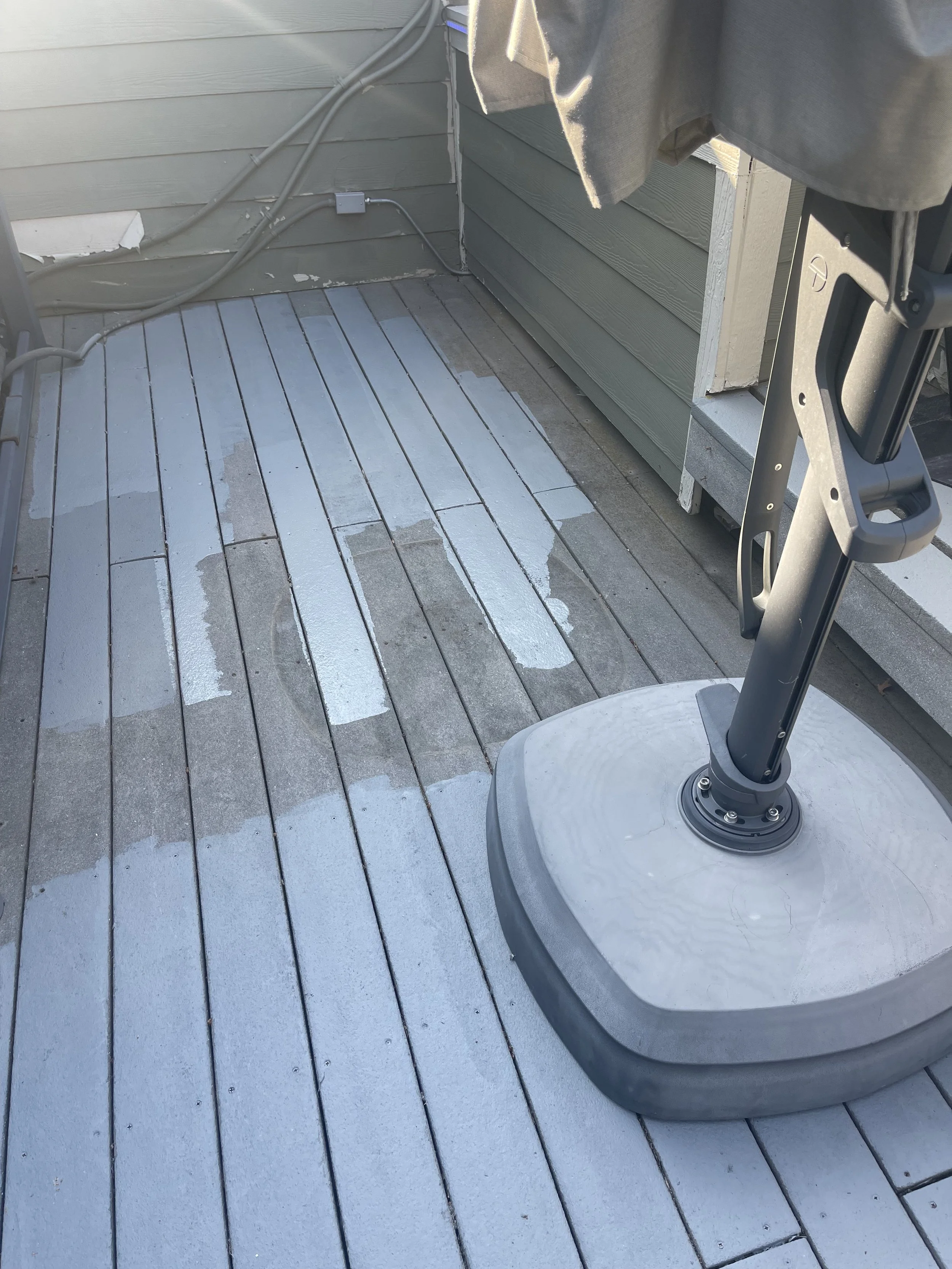A patio with wooden floorboards, some of which are painted white, as part of a painting project. An umbrella stand with a folded umbrella is in the foreground, and a section of a house's exterior wall with electrical wiring is visible in the backgrou
