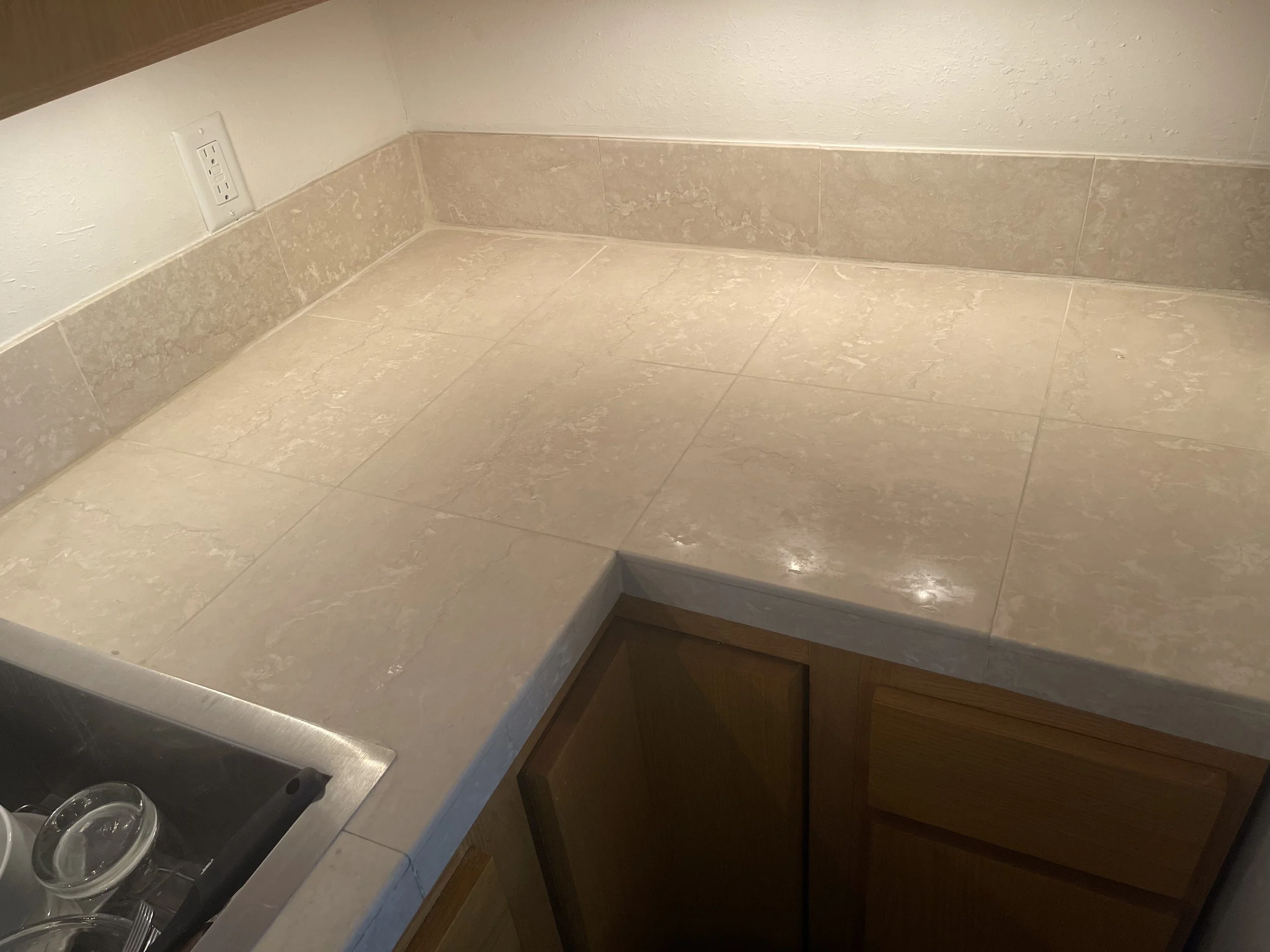 Kitchen countertop with beige tiles, a brown cabinet, and a nearby kitchen sink with glassware.
