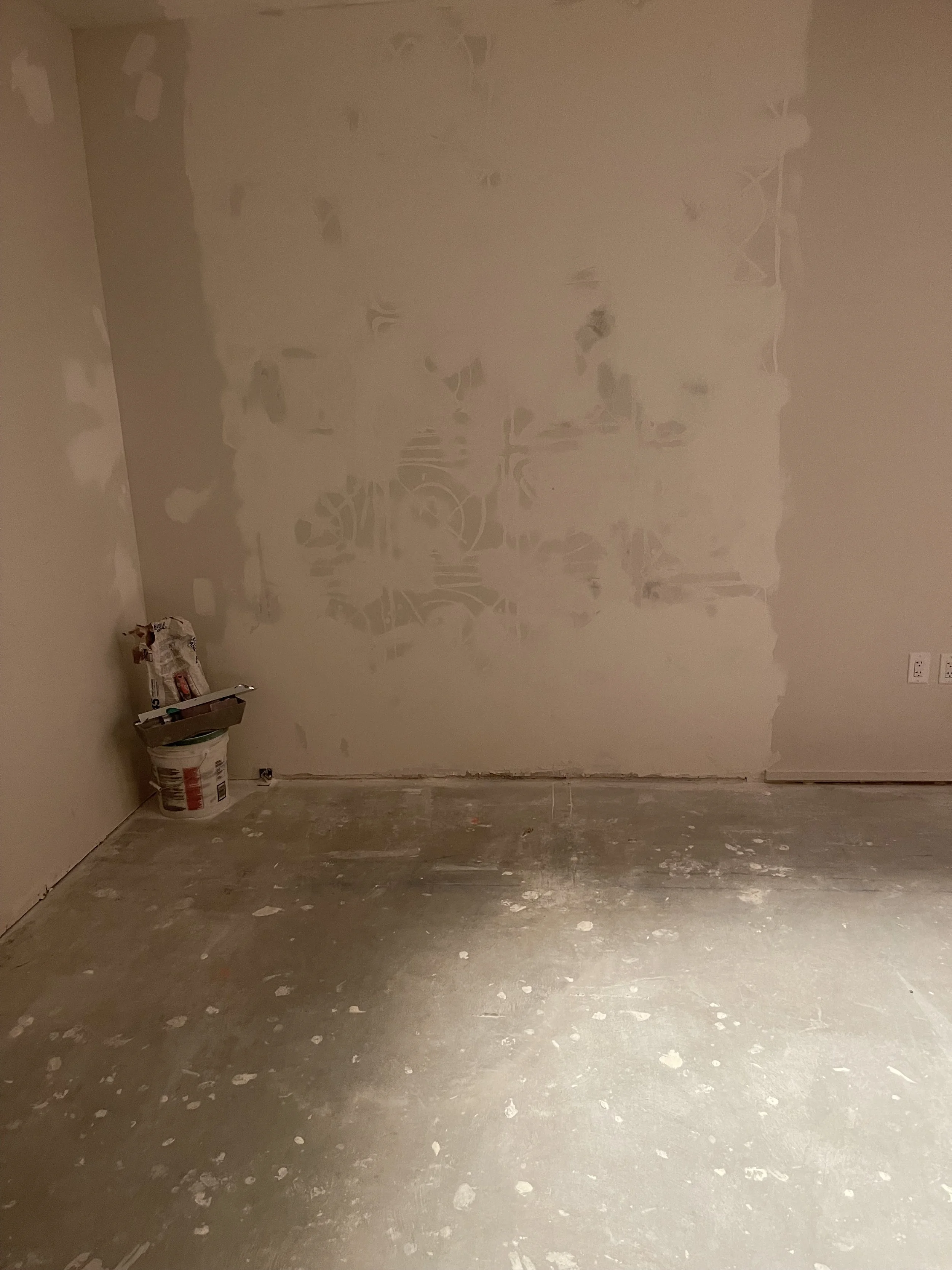 Interior room under renovation with partially finished drywall and a concrete floor, with construction supplies in the corner.