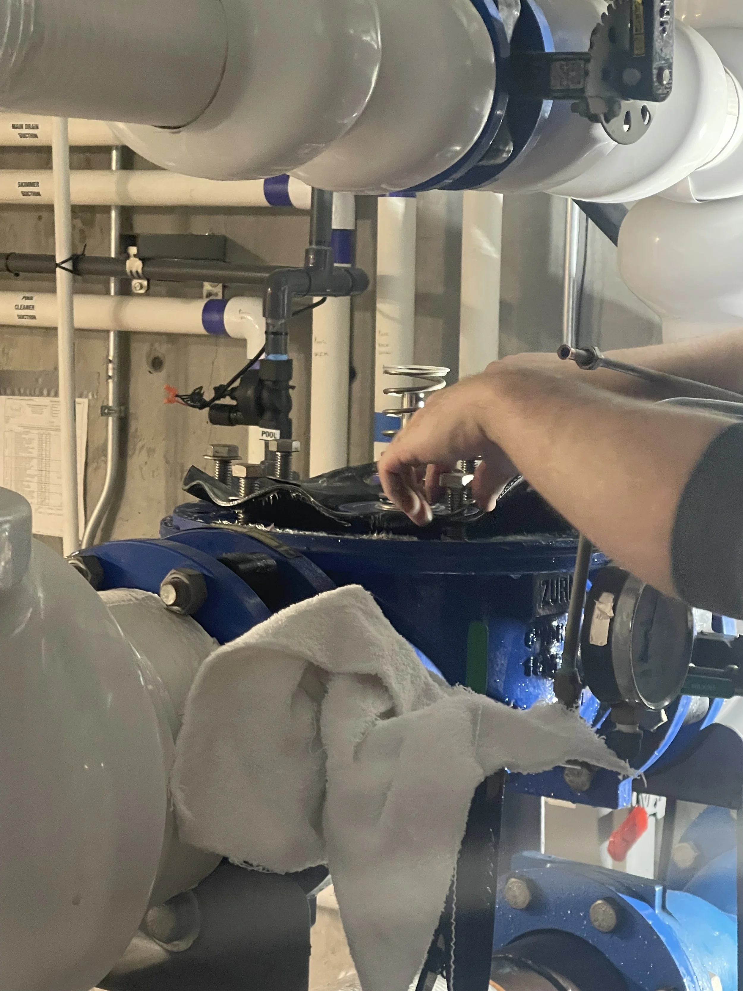 A person working on machinery in an industrial setting, with pipes, gauges, and tools visible, focusing on assembling or repairing equipment.