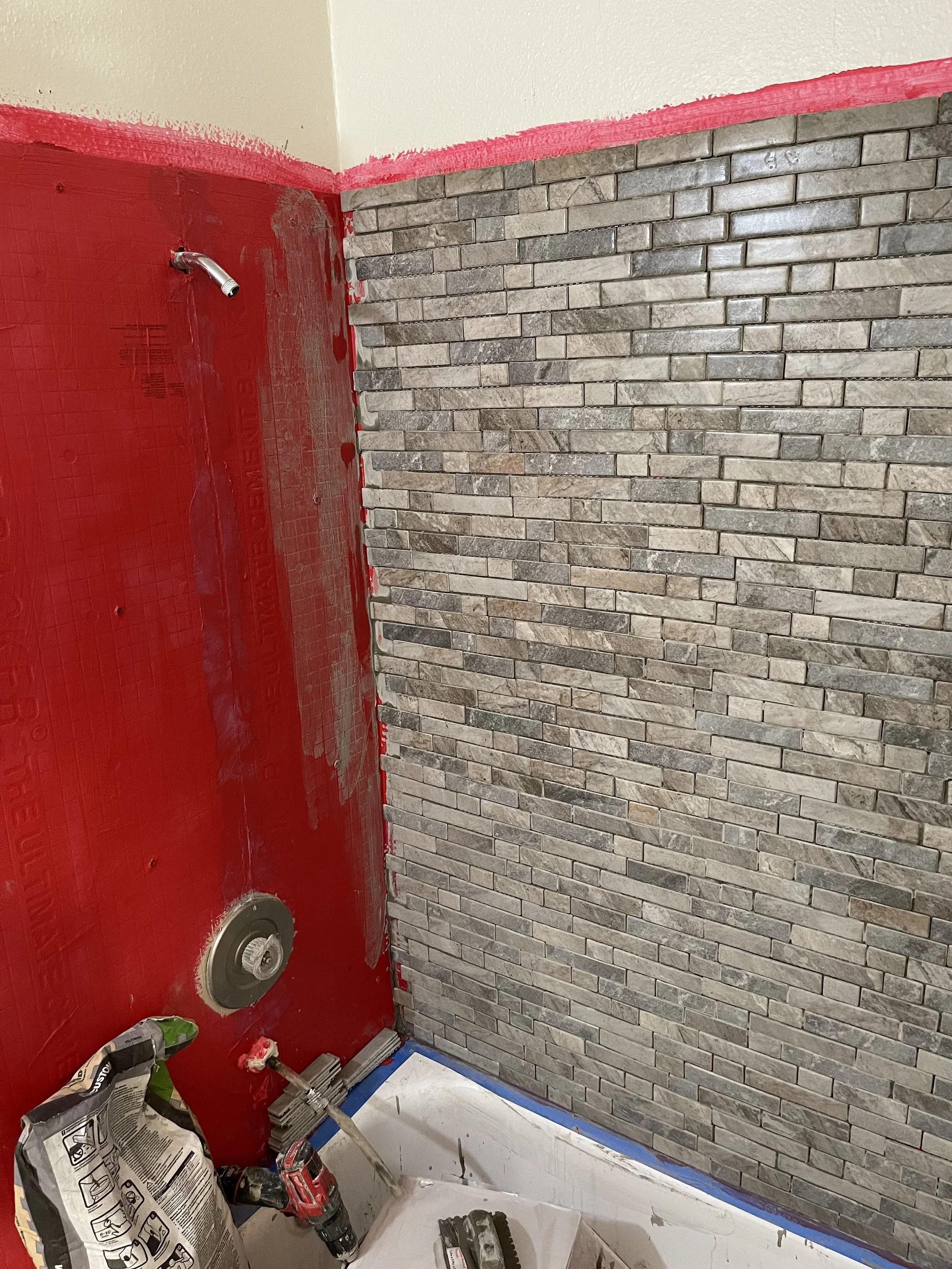 Bathroom shower area under construction with gray brick wall on the right, red waterproofing on the left, and a showerhead installed. Tools and materials are on the floor.