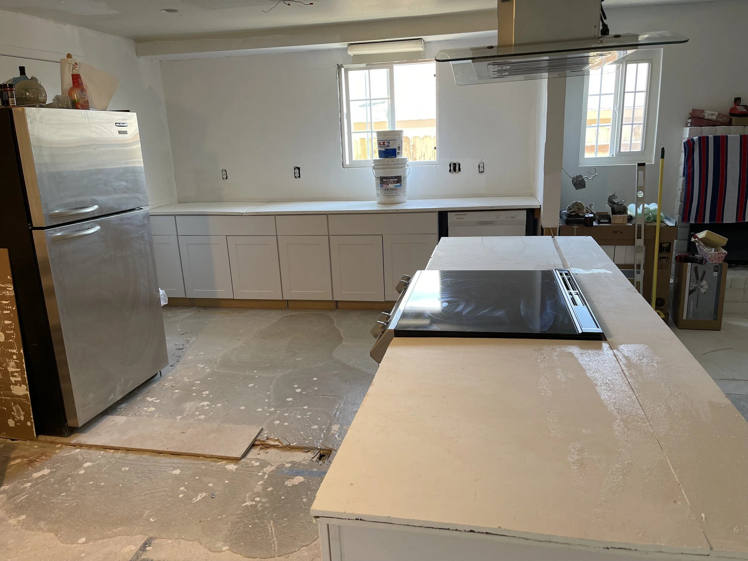 Kitchen under renovation with white cabinets, stainless steel refrigerator, and a black electric cooktop on a white island, construction materials, and tools scattered around.