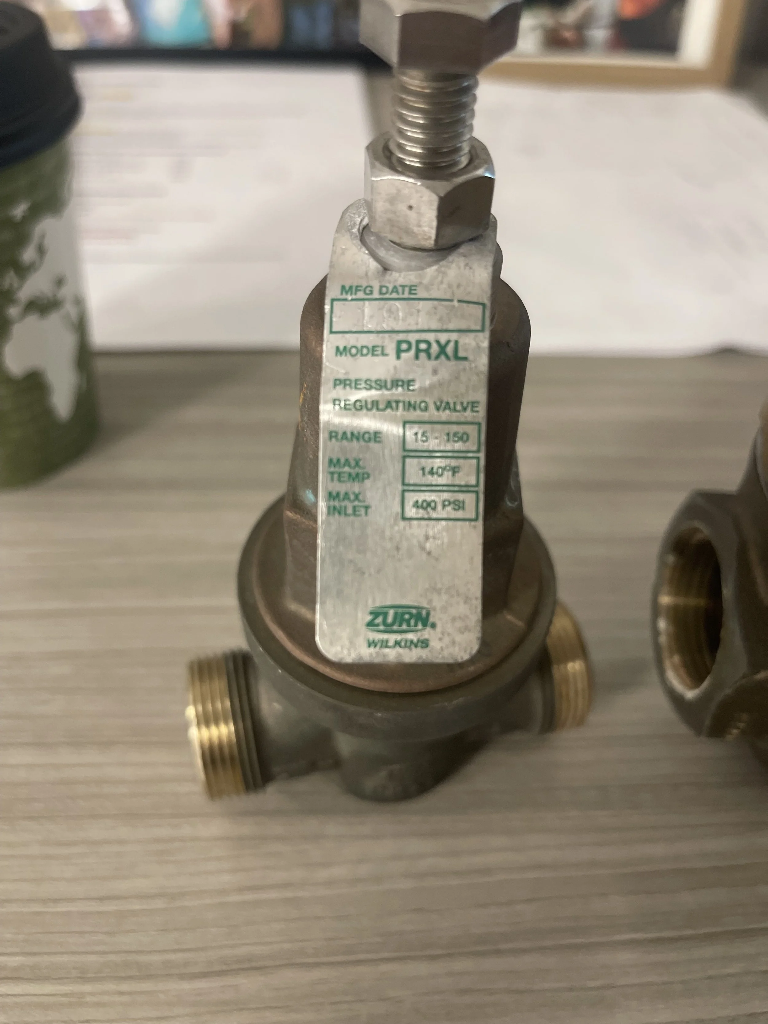A pressure regulating valve with a metal tag displaying specifications such as model PRXL, pressure range 15-150 PSI, max temperature 140°F, and max inlet pressure 400 PSI, installed on a wooden surface with other plumbing components and a green cont