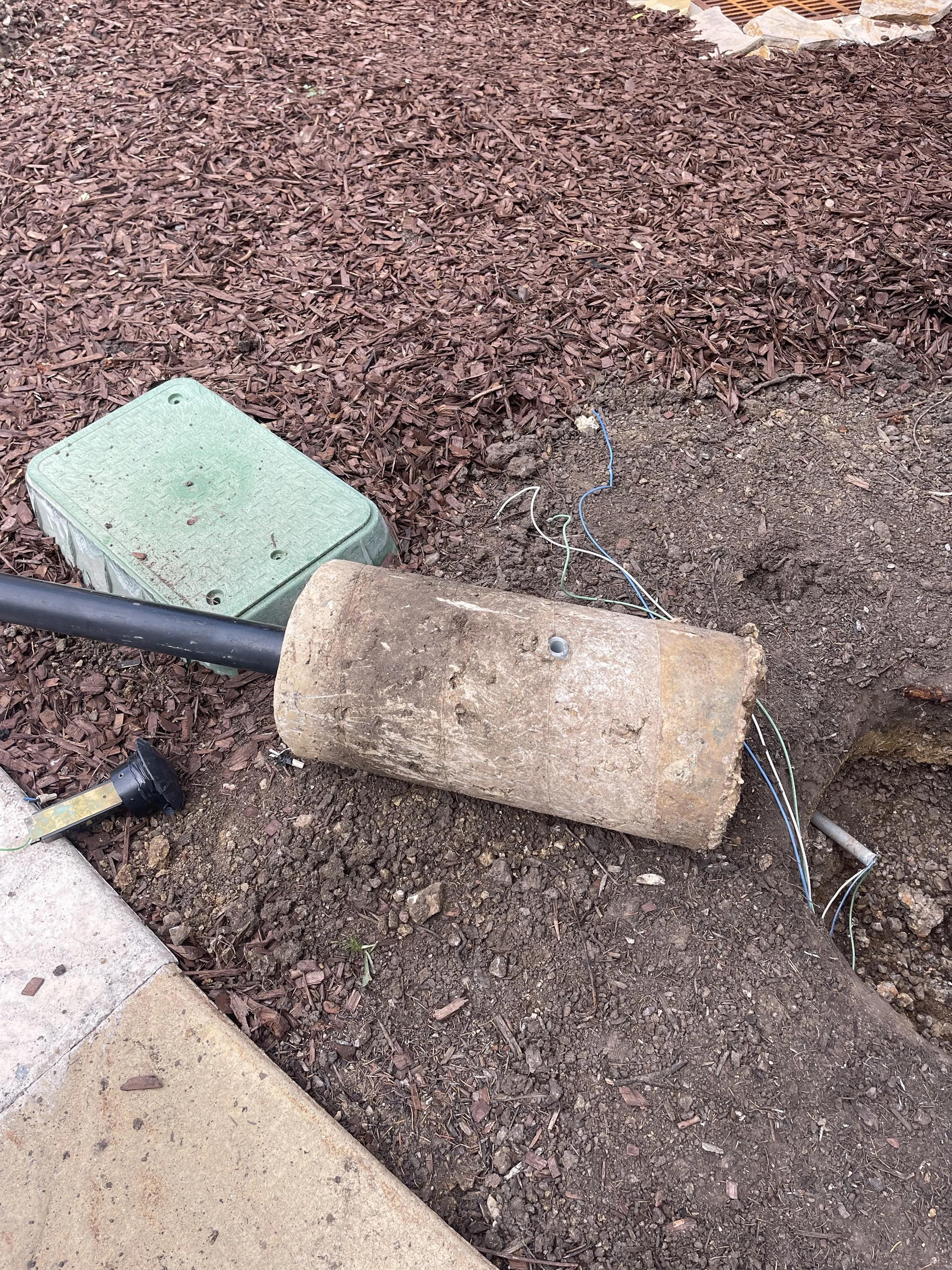 An exposed electrical conduit or pipe, partially buried in ground, with electrical wires coming out. Nearby is an electrical box and some mulch on the ground.