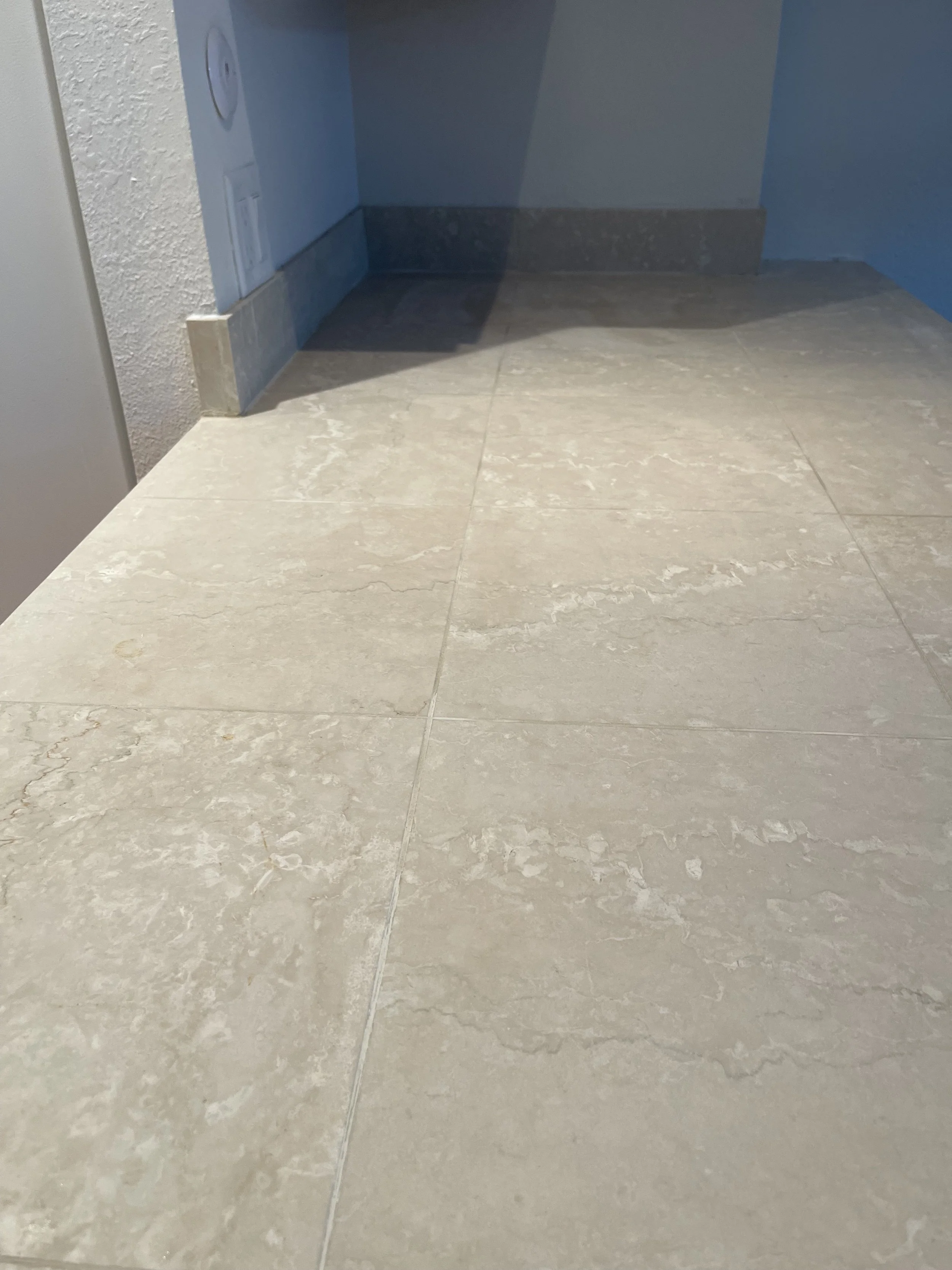Close-up of beige marble floor tiles with grout lines, part of a light-colored wall, an electrical panel and outlets, and a darker blue wall in the background.