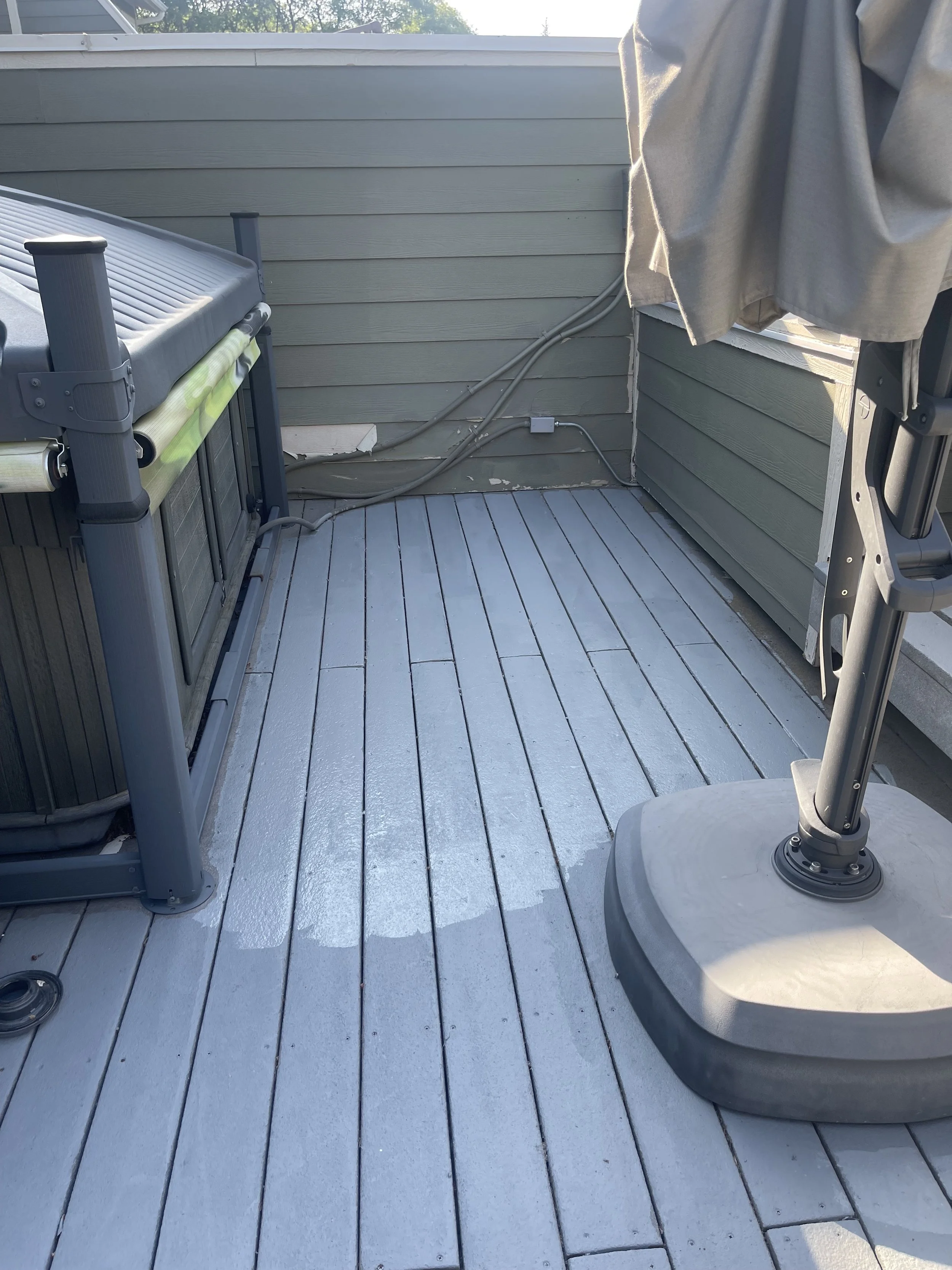 A small outdoor deck featuring a hot tub, a patio umbrella stand, and some electrical cords along the wall, with a gray wooden floor and green siding.