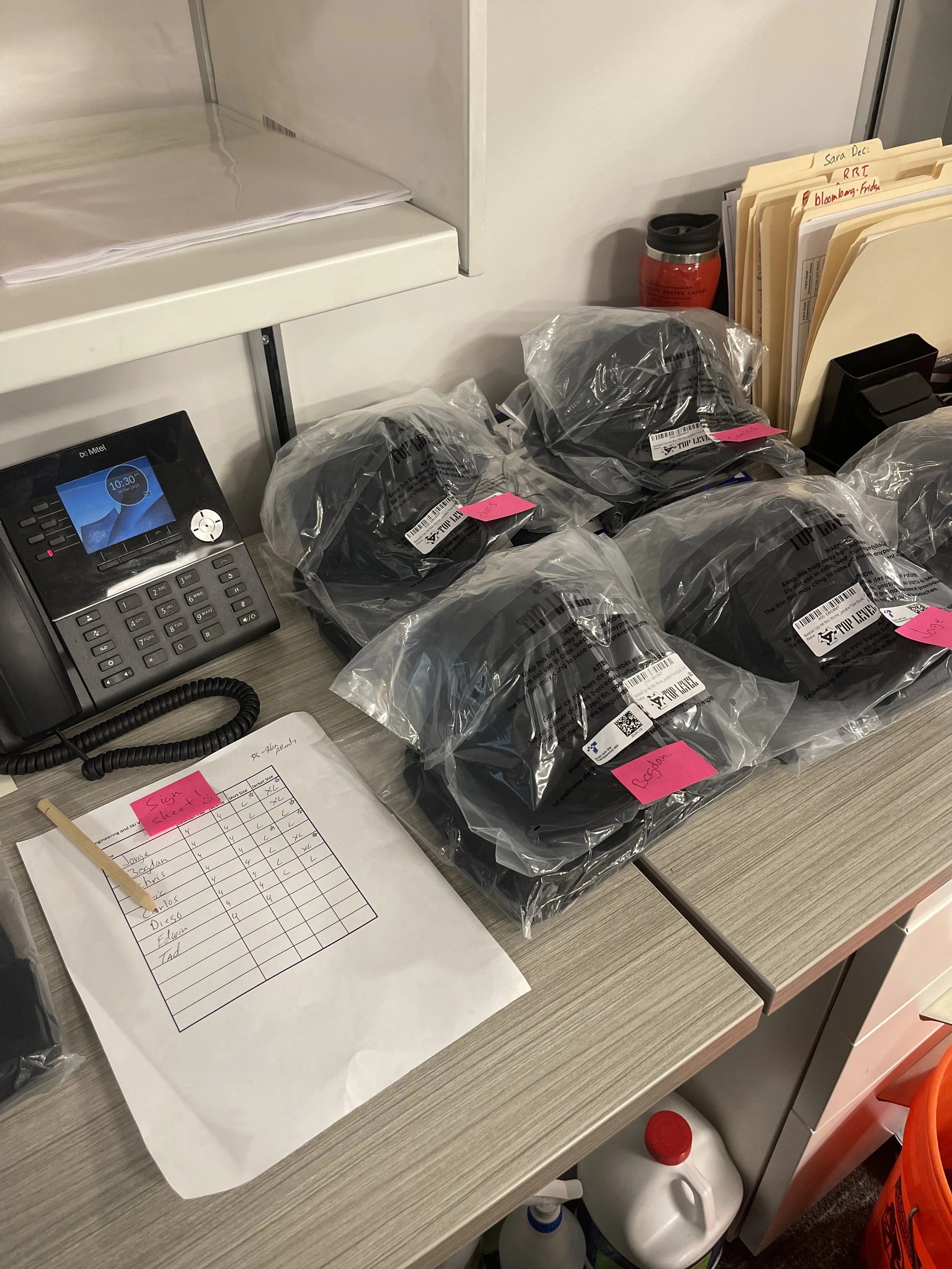 Multiple sealed bags containing black fabric hats or garments are placed on a gray office desk. There is a telephone, a sheet of paper with a handwritten chart, and some folders and files on the desk. A red and black travel mug and a monitor are also