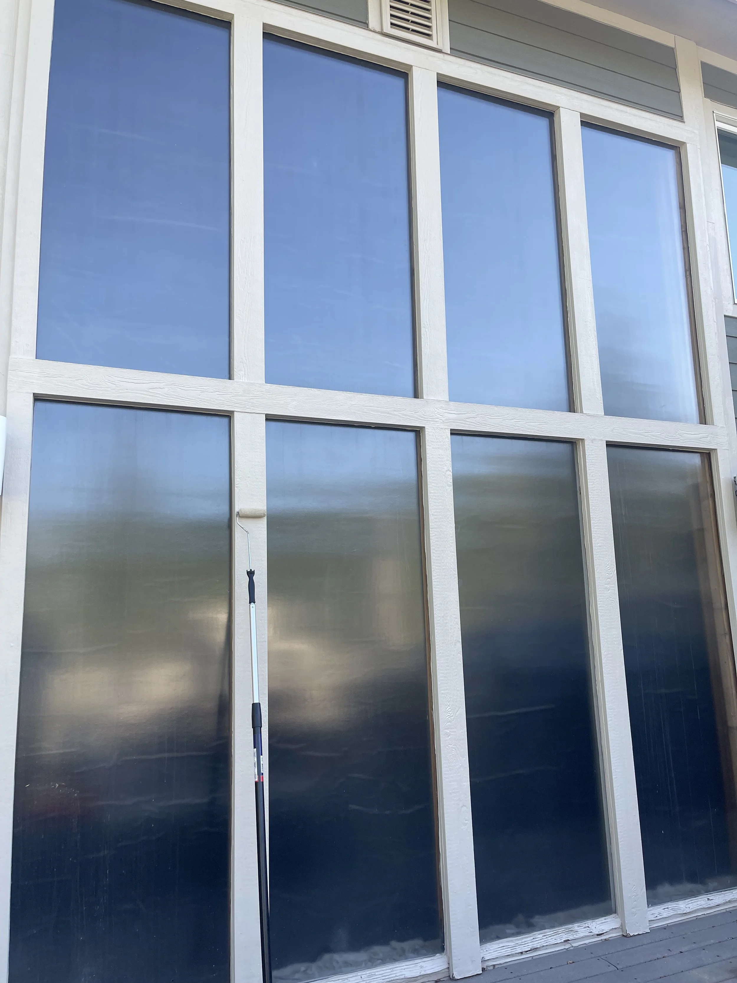 Large windowed door with multiple glass panels framed in white, with some panels covered and others reflecting the sky, with a black-handled rolling act or screen in front.