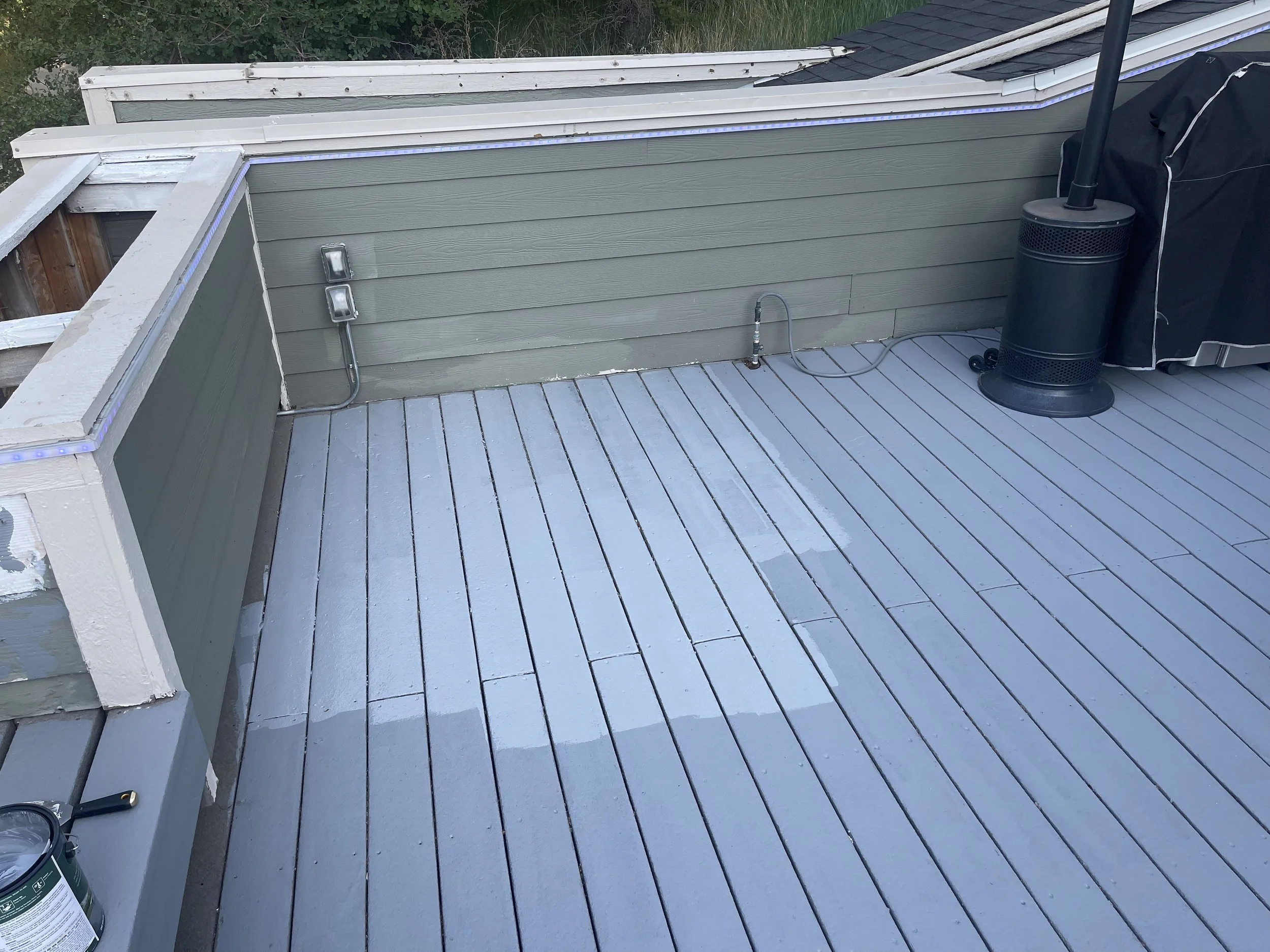 A deck with new gray painted wooden planks, a green wood-paneled wall with electrical outlets, a black outdoor heater, and a covered grill.