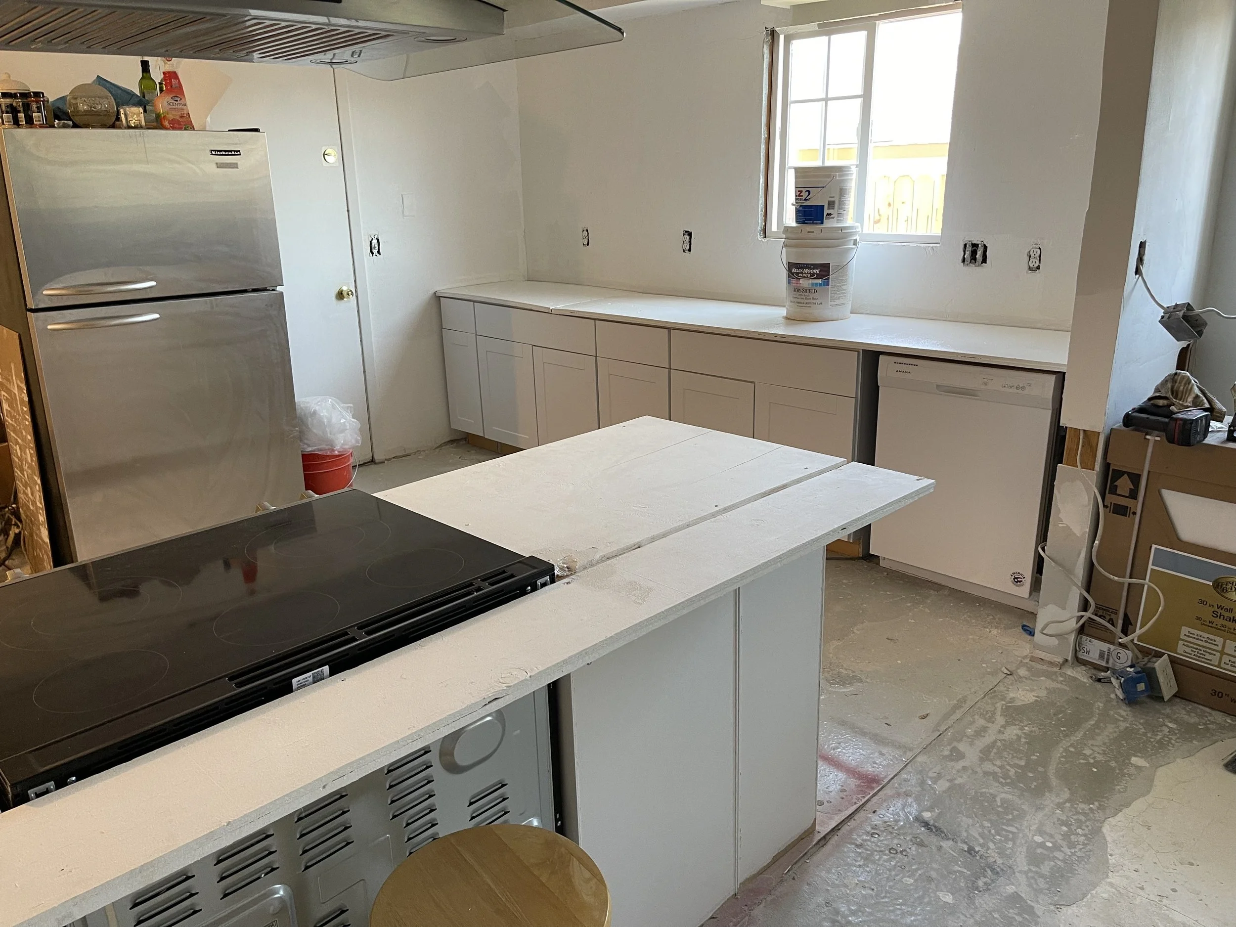 Kitchen under renovation with white cabinets, a black electric stovetop, a stainless steel refrigerator, a window with sunlight, and construction supplies scattered on the floor.