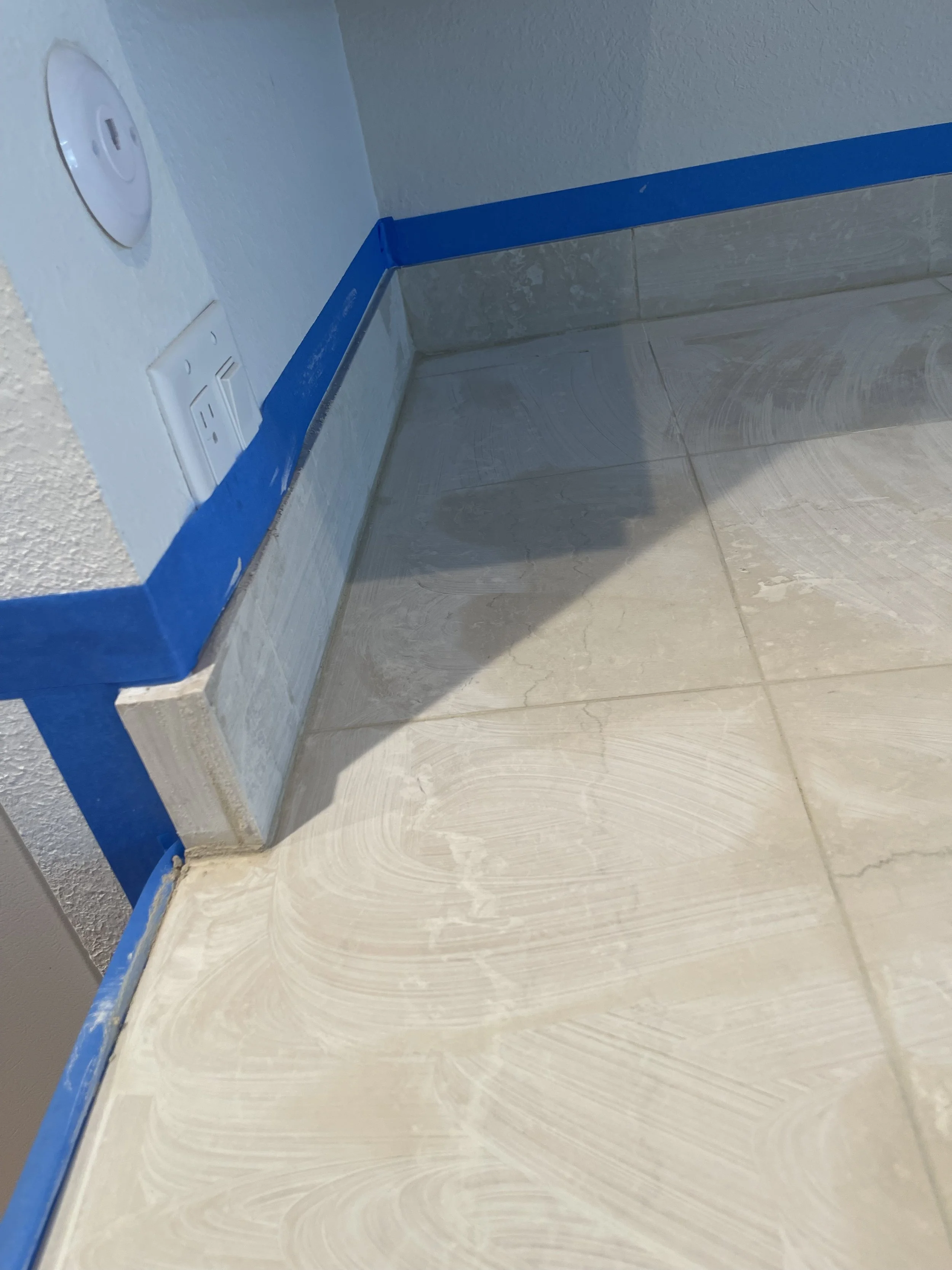 Part of a wall and floor corner being prepared for painting, with blue painter's tape along the baseboard, electrical outlets, and a wall plate visible.