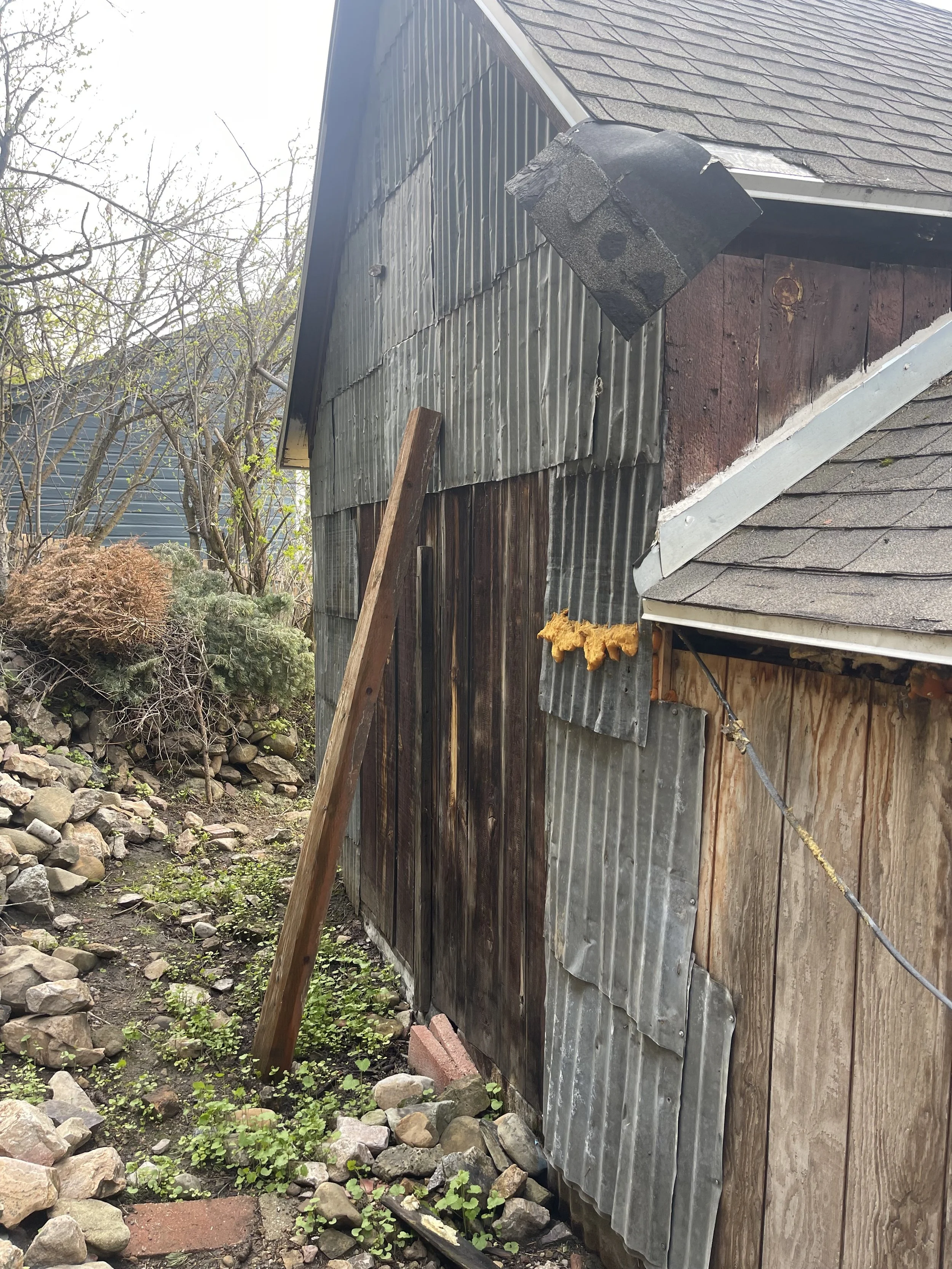 Side of a building with mismatched siding, including wood, corrugated metal, and asphalt shingles with a metal flashing. There is a wooden board propped against the building and some rocks and plants on the ground.