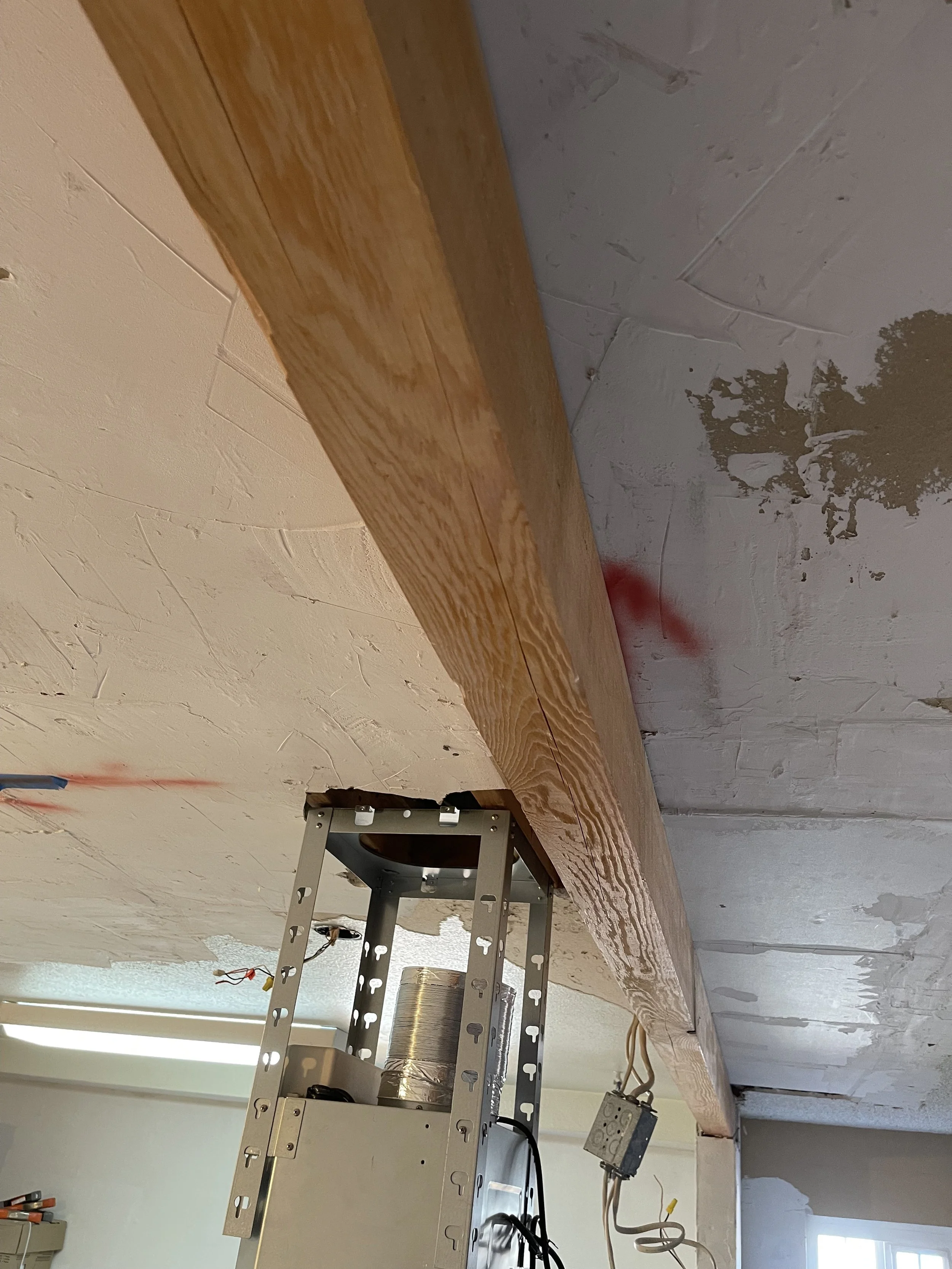 Close-up view of a wooden beam installed under a ceiling with drywall and patches, with construction tools and wiring visible.