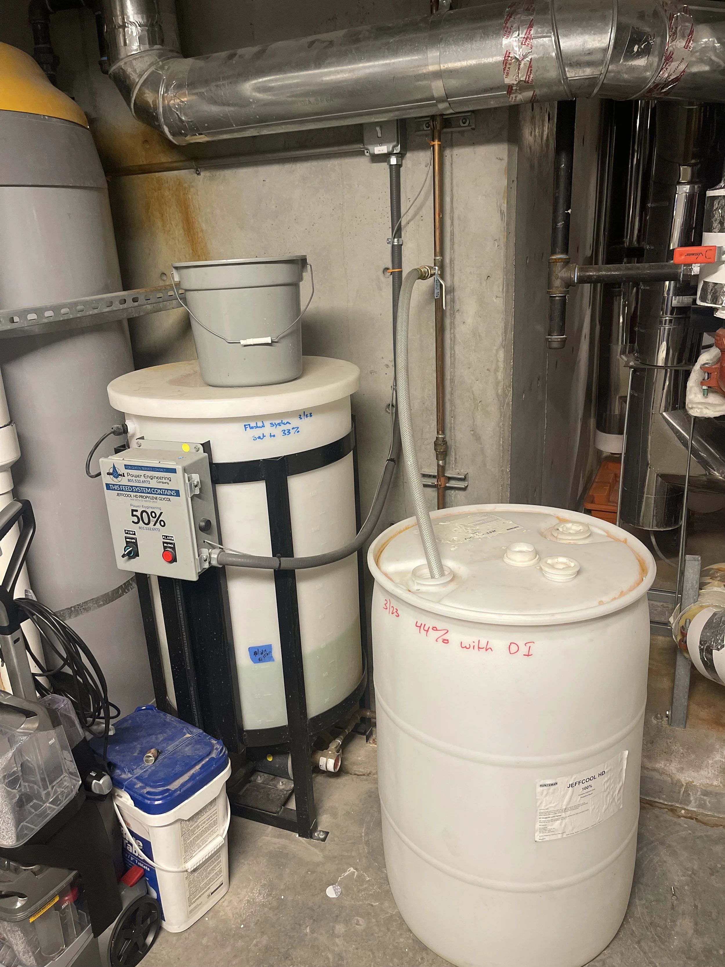 A basement utility room with a large white fermentation tank, a white plastic barrel, and various pipes and equipment used for brewing or fermentation processes.