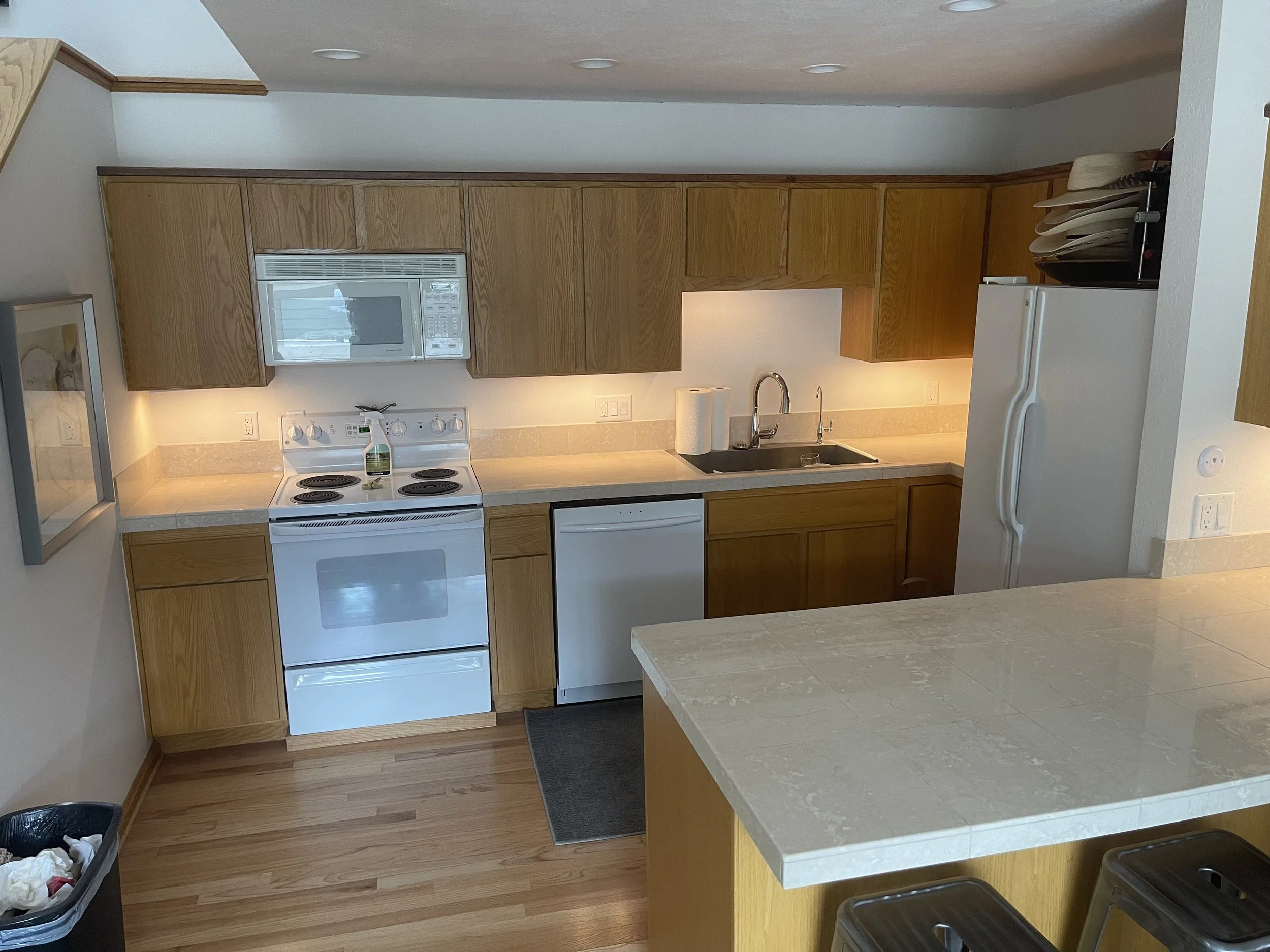 View of a kitchen with wooden cabinets, a white stove, microwave, dishwasher, and refrigerator. There is a sink with a faucet, paper towel rolls, and a countertop. Some dishes are stacked on top of the refrigerator, and a small trash can is on the fl