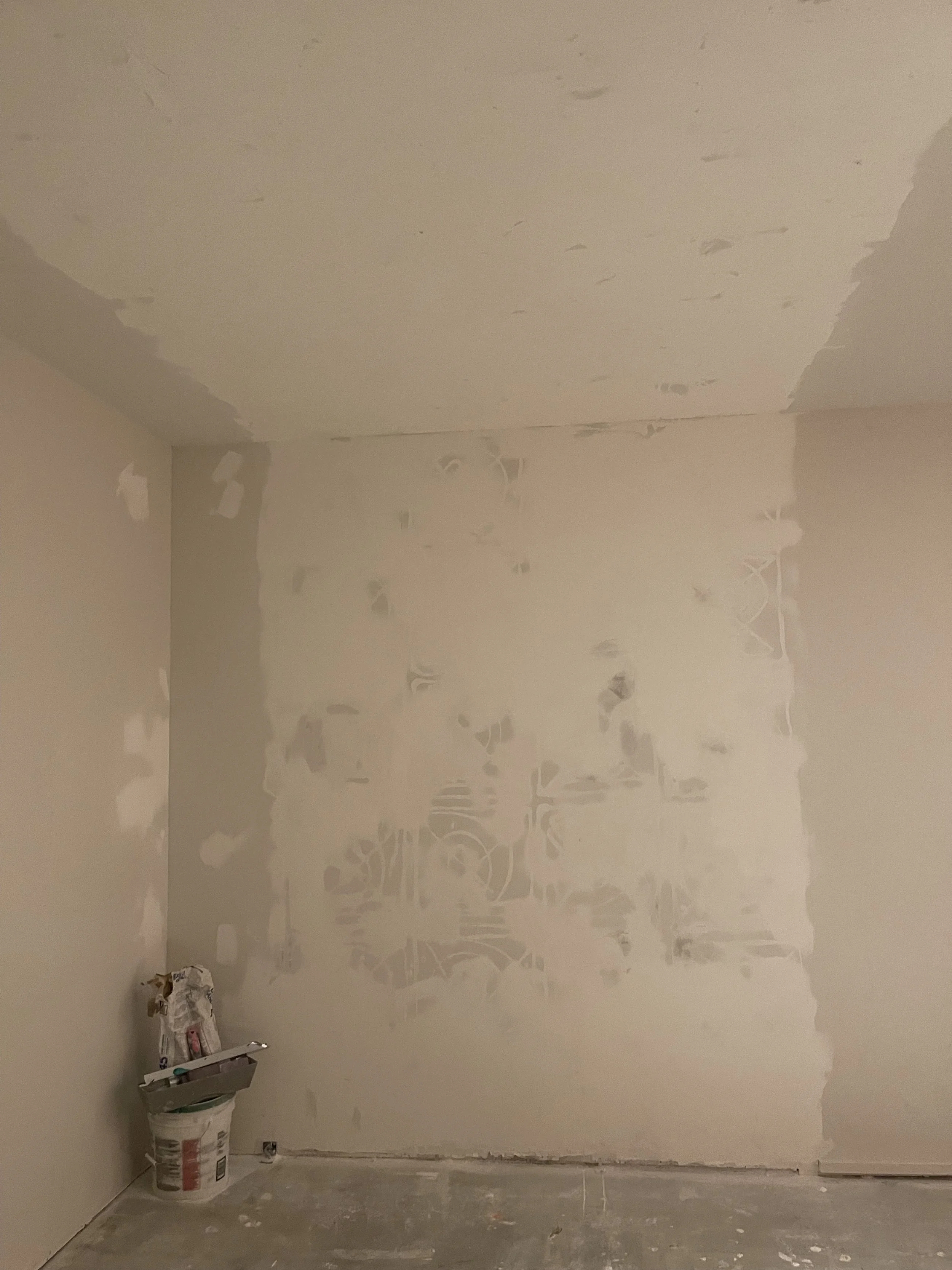 A room under construction with drywall installation in progress. The walls and ceiling are partially taped and mudded, and there is a bucket of joint compound with tools in the corner.